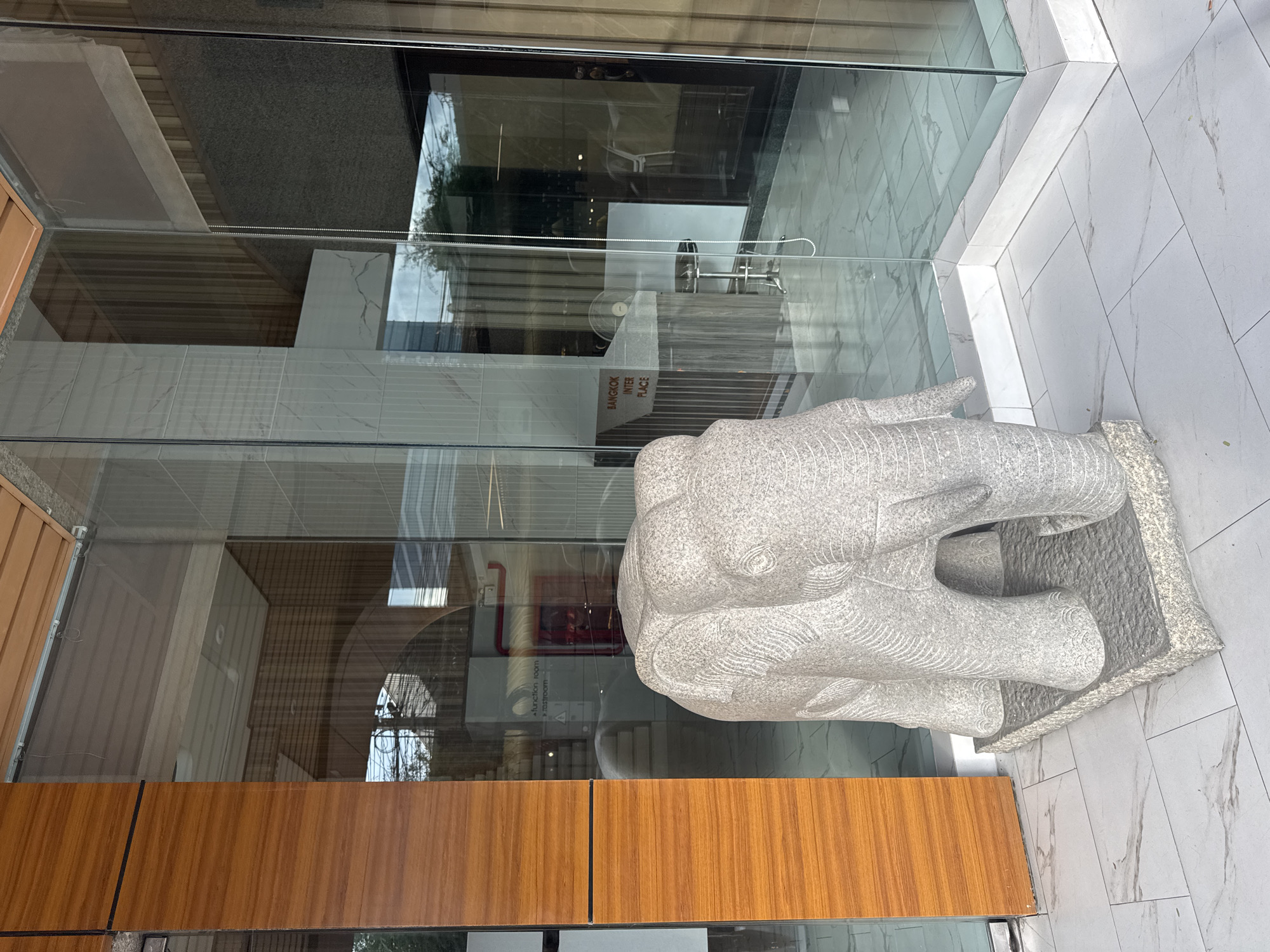 The image, presented in a portrait orientation, captures a static scene featuring a large, light-gray, granular stone elephant statue positioned outdoors, adjacent to a modern glass-fronted building. The elephant, detailed with carved folds on its ears and trunk, rests on a rectangular stone base.
The glass facade creates a complex visual, simultaneously revealing the building's interior and reflecting its exterior surroundings. Inside the building, visible through the glass, is a reception-like area with what appears to be a dark-colored counter or desk, topped by a white electric fan. A vertical sign within the building clearly reads "BANGKOK INTER PLACE," serving as a prominent location cue consistent with the provided context of Bangkok, Thailand. Further inside, obscured reflections hint at "function room" and "washroom" signage, along with a red object that might be a fire alarm or extinguisher.
Reflected in the glass are elements of the outdoor environment, including parts of green trees or foliage, and what seems to be a bright, possibly overcast, daytime sky. The ground outside and the floor inside the building are covered in large, light-colored, marble-patterned tiles. To the immediate right of the elephant, a geometrically patterned wall section is also tiled in a similar style. A horizontal band of warm-toned wooden paneling runs across the lower portion of the image, likely part of an exterior wall or planter box. No people or specific activities are depicted, emphasizing the architectural and sculptural elements of the scene.
