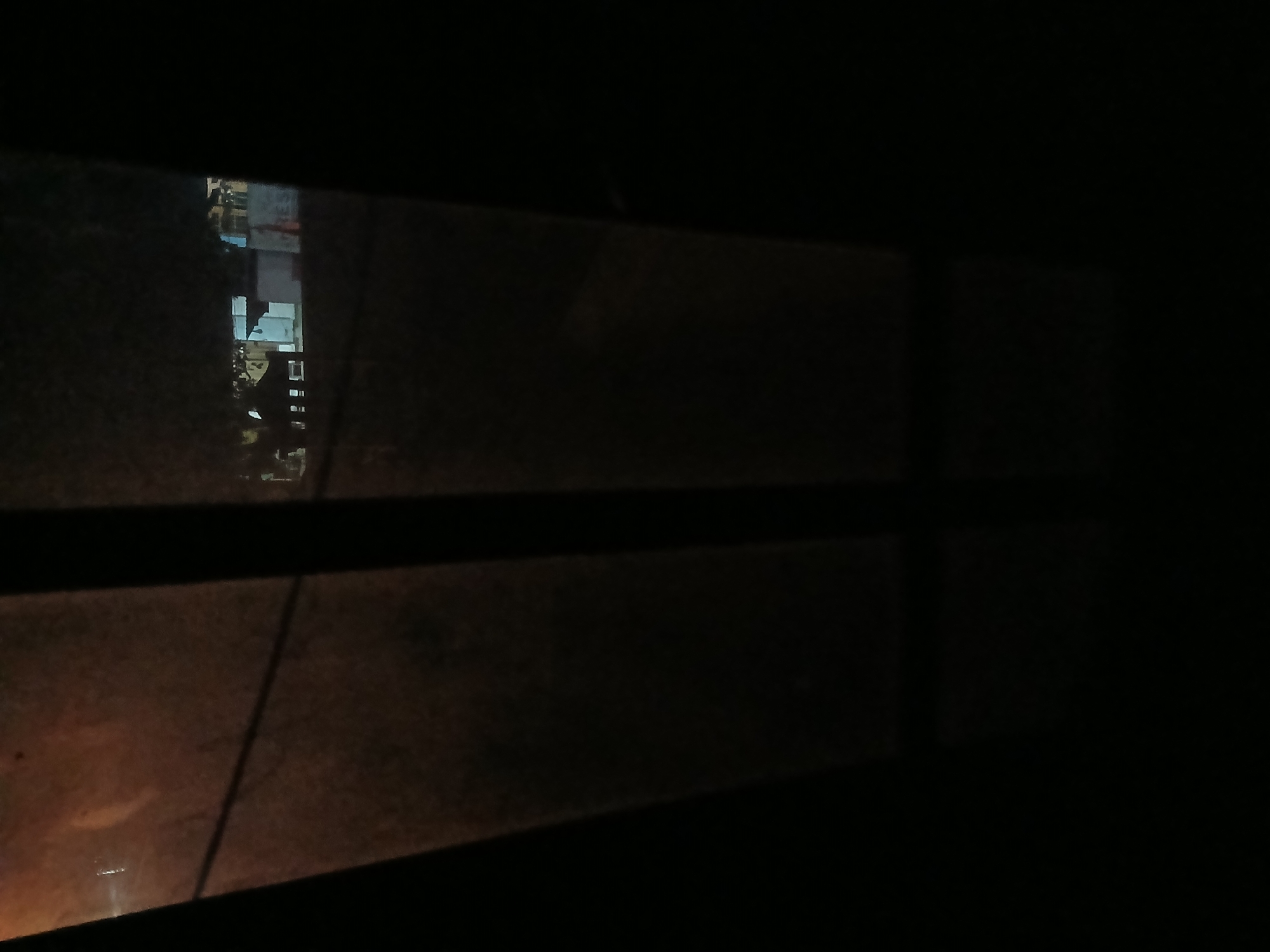 The image shows a view through a window at night. In the background, through the slightly dirty glass, there are buildings with lights on. The setting appears to be urban or suburban. The foreground is dominated by the dark interior of the room from which the photo is taken, with the window frame creating a division in the view. There are no people visible in the image.
