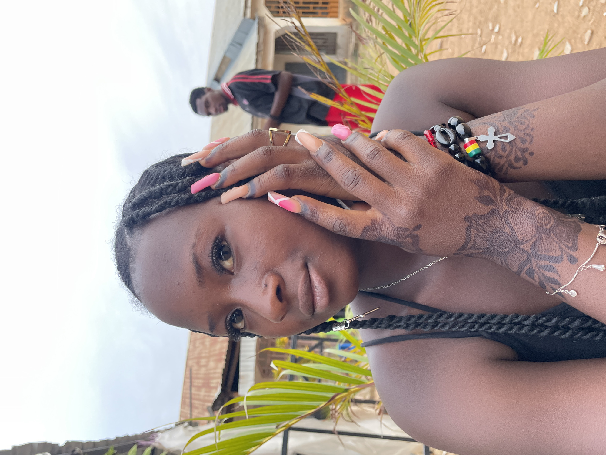 A young Black woman is the primary subject of this portrait-style photograph. She has dark brown skin, dark eyes, a nose piercing, and her hair is styled in dark braids with silver and gold accents. Her hands are close to her face, displaying a gold geometric ring on her finger and long, manicured nails with a pink and beige color scheme. On her right arm, visible tattoos, a silver bracelet, and a black beaded bracelet with a silver cross charm and the colors of the Ghanaian flag are noticeable.
Behind her, slightly blurred, is a man in a black and red athletic outfit. The background suggests an outdoor environment, possibly in Jos, Nigeria, with elements like a building with an exposed brick roof and a sandy hill visible, as well as green vegetation. The sky is overcast. The image is slightly rotated to the left, as if the camera was held at a slight angle.