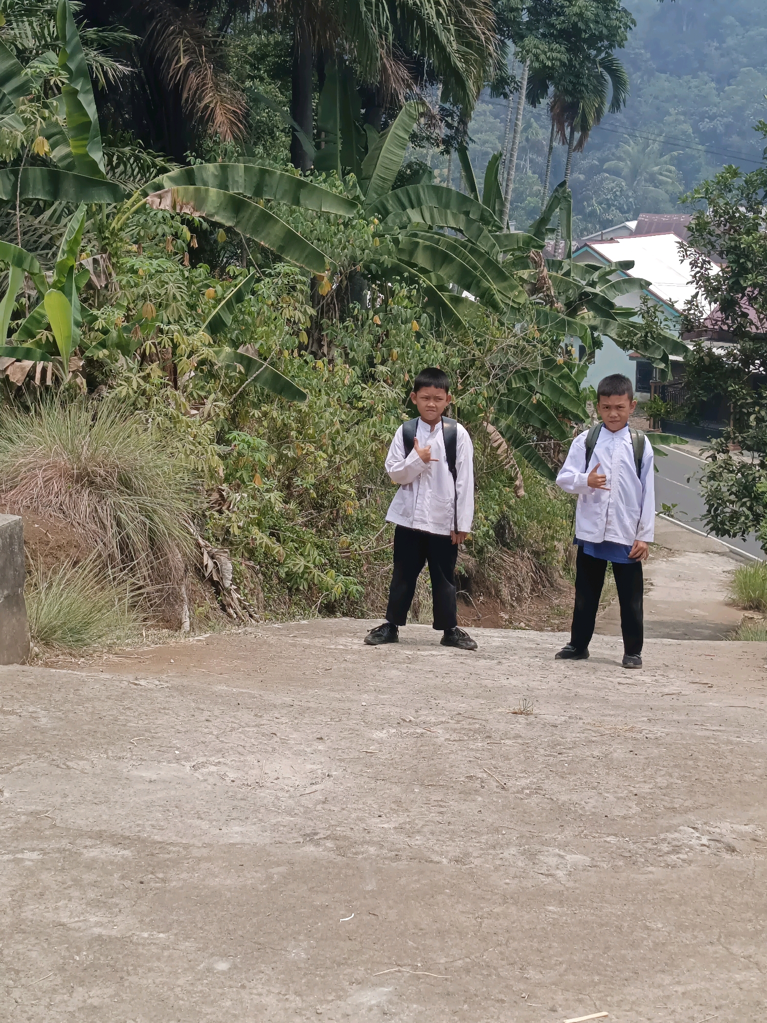 Two young boys are standing on a concrete pathway. The boy on the left is wearing a white long-sleeved shirt and black pants, with a black backpack. He has his right hand raised with his thumb and index finger extended in a gesture. The boy on the right is also wearing a white long-sleeved shirt and black pants, with a black backpack. He has his right hand raised in a similar gesture. They are standing in front of lush green vegetation, including banana trees and other foliage. In the background, there are houses with white roofs and a winding road. The scene appears to be in a hilly, tropical environment, consistent with the landscape of Taba Tinggi, Indonesia. The overall lighting suggests it is daytime, likely overcast or in the shade.