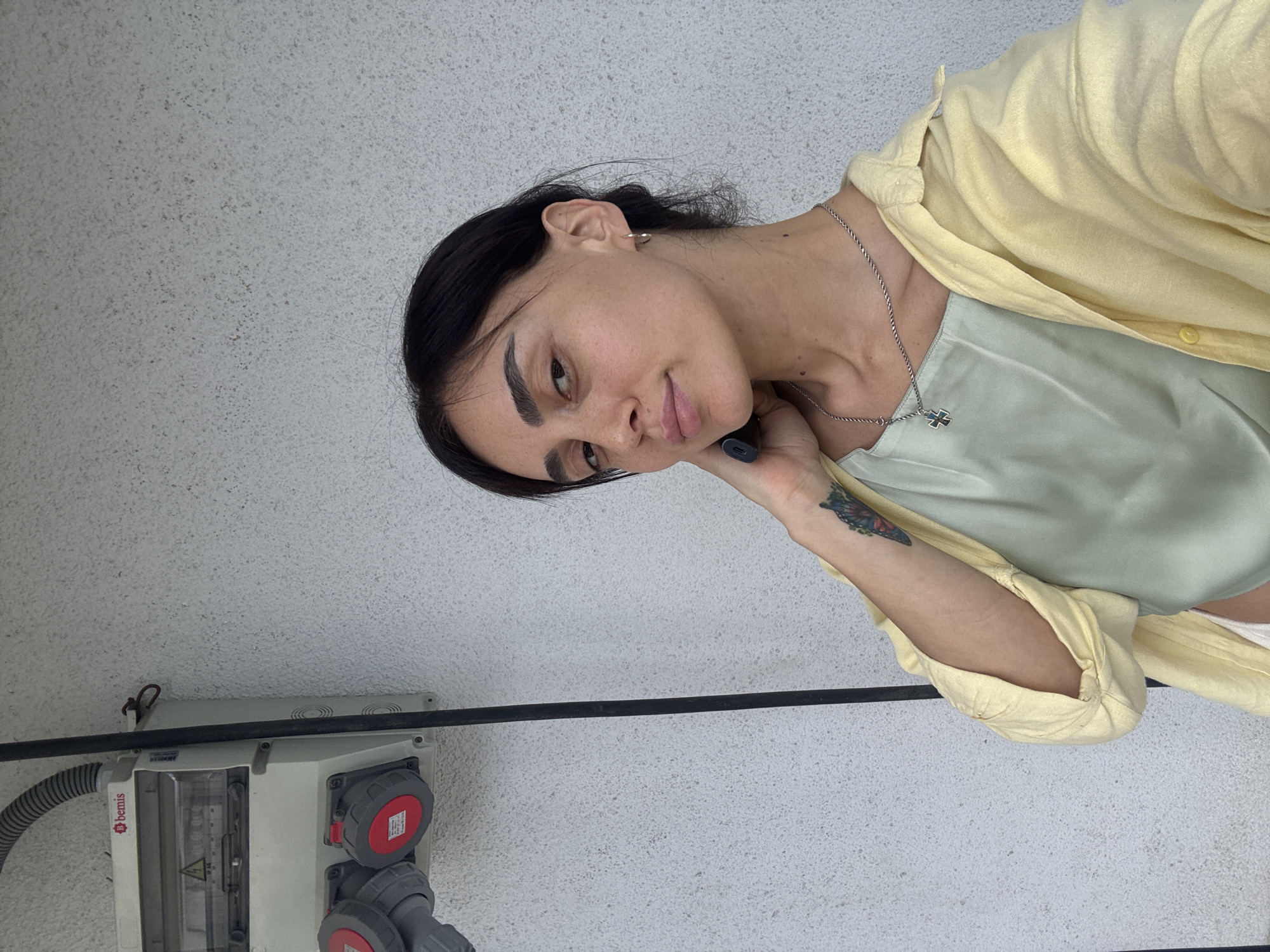 A young woman with dark hair and dark eyes poses for a selfie against a textured white wall. She is wearing a pale yellow open shirt over a light green slip dress, and a silver chain with a cross pendant. A tattoo of a butterfly is visible on her left forearm. To the left of the woman is an electrical panel with red and gray outlets. The image appears to have been taken outdoors.