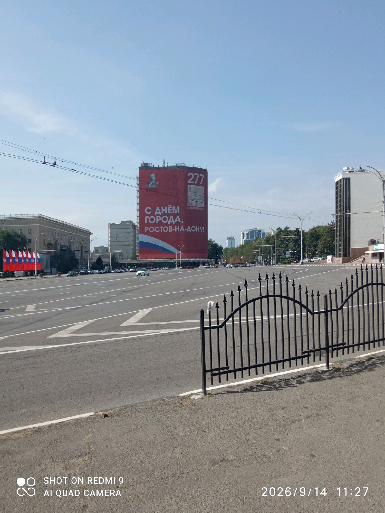 The image depicts a wide, empty urban intersection on a clear day. Dominating the background is a large, red banner on the side of a building, featuring a portrait of a man, the number "277," and Russian text that translates to "Happy City Day, Rostov-on-Don!" A Russian flag motif is also visible. To the left of the banner, a long, light-colored building with many windows lines the street, and several flags are displayed on poles in front of it. To the right of the banner, a modern, white building with a glass facade stands tall. The foreground features an asphalt road with distinct white lane markings. A black, wrought-iron fence runs along the edge of the road, separating it from a paved area. The sky is bright blue with a few wispy clouds. The overall scene suggests a civic celebration or event in Rostov-on-Don, Russia.