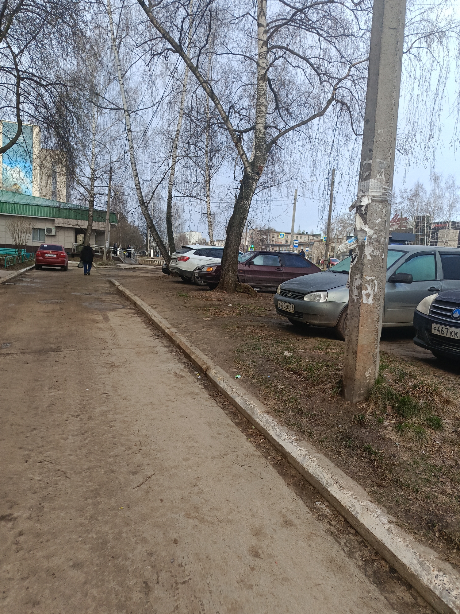 The image shows a street scene in what appears to be a residential or mixed-use area, potentially in Chaikovsky, Russia, based on the visible license plates. The foreground features a concrete curb and a somewhat dirty paved area, likely a sidewalk or pedestrian path. To the right of the sidewalk, a dirt area is visible with sparse grass and parked cars. Several cars are parked along the side of the road, with the front car partially obscured by a utility pole covered in peeling paper. Further down the road, more parked cars can be seen, along with a person walking away from the camera. In the background, there are buildings, trees, and utility poles. The trees are mostly bare, suggesting it is early spring or late fall. The sky is overcast. The overall impression is of a typical street scene in a Russian town, possibly Chaikovsky, based on the license plates on the vehicles. The location appears to be a regular street with parked cars on the side, and a sidewalk for pedestrians.