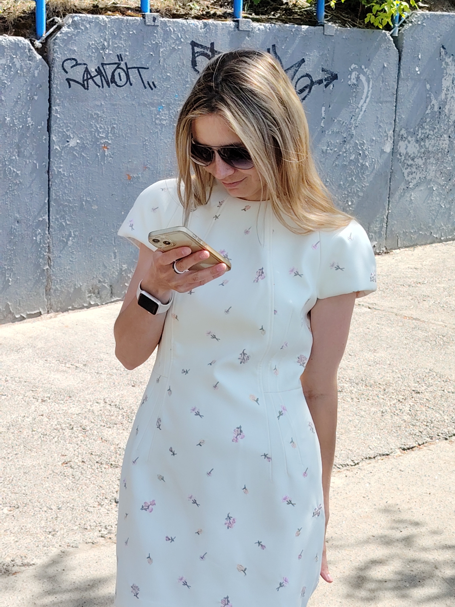 A woman stands outside in Chaikovsky, Russia, looking at her cell phone. She is wearing sunglasses and a white dress with small pink flower patterns, as well as an Apple watch. Behind her is a gray concrete wall with graffiti. A blue metal fence is visible on top of the wall. The ground appears to be paved with asphalt or concrete.