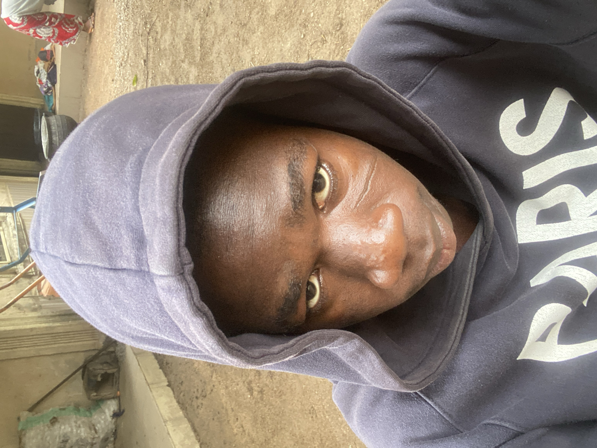 An outdoor, eye-level portrait shot in Mararaba, Nigeria, features a young individual with dark skin, wearing a dark blue hooded sweatshirt with "PARIS" in white lettering on the chest. The subject looks directly into the camera with a stern expression. Distinctive linear facial scarifications are visible on the left cheekbone. The hood of the sweatshirt is pulled up, framing the face. The skin on the face appears shiny.
The background consists of a dusty, light brown gravel and dirt ground. To the left, a light-colored building with vertical fluting on its wall is visible, fronted by a raised concrete platform. On this platform, a large, green and white striped sack rests, with a dark, box-like object behind it and a thin rod leaning against the wall.
To the right, another section of a similar building is visible. A dark pot or container sits on a raised platform. Further right, a second person is partially visible from the waist down, wearing a white top and a red and white patterned skirt or wrapper, with light-colored sandals. This individual is bent over, facing away from the camera, near other assorted colorful fabrics or items on the platform. The scene is illuminated by natural daylight.