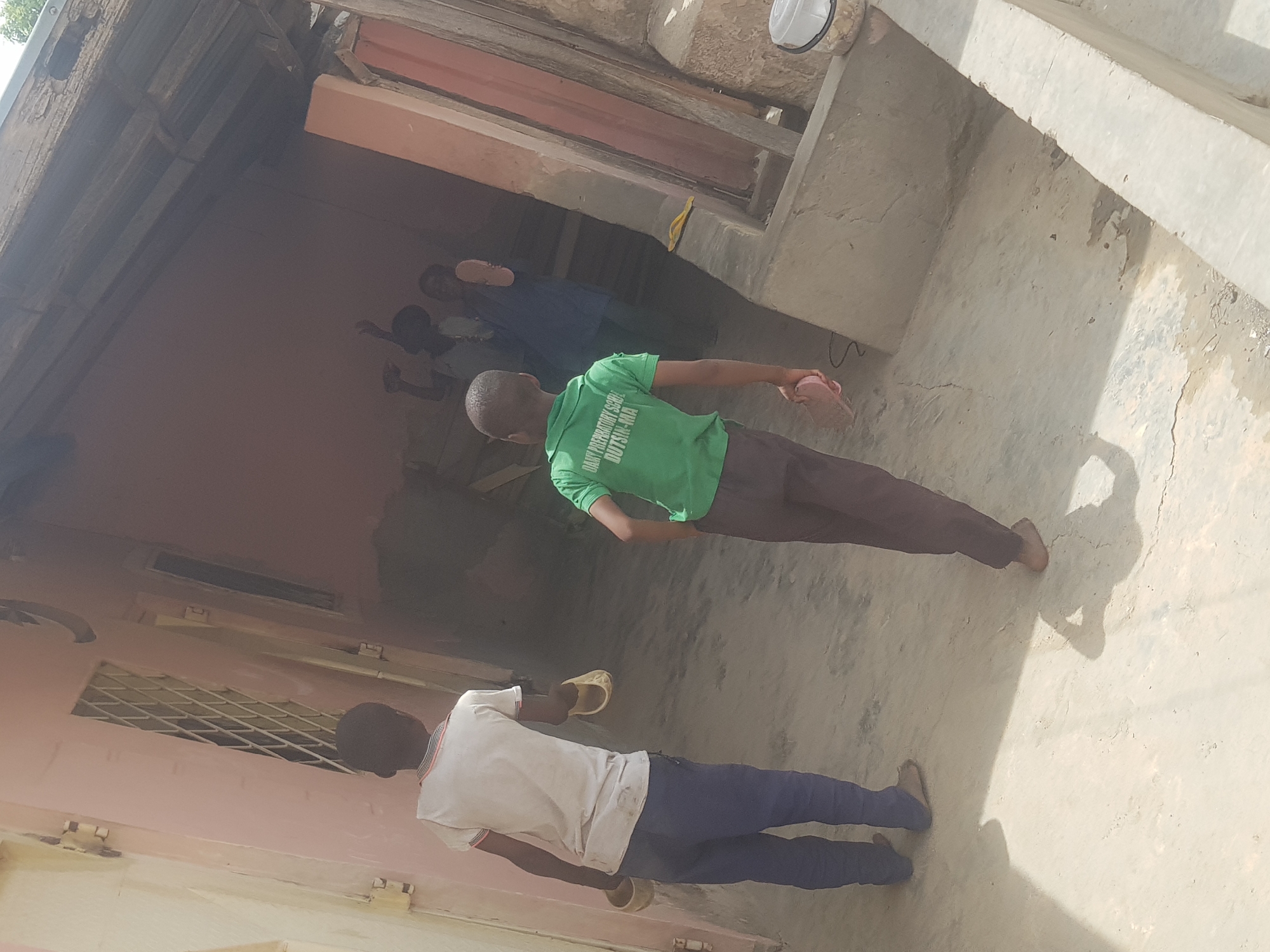 The image shows an outdoor scene in Dutsinma B, Nigeria. Two young men are visible in the foreground. One wears a white shirt and blue pants, holding a yellow sandal. The other wears a green shirt with text and brown pants, also holding a sandal. In the background, three other individuals are partially visible inside a building. The surrounding environment consists of buildings with light-colored walls and concrete ground.