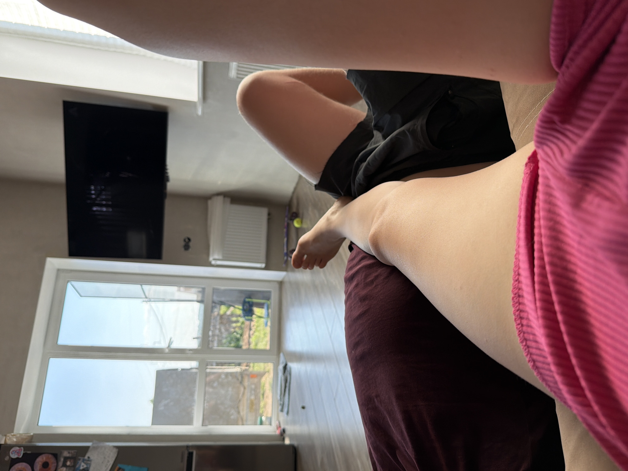 The image shows an indoor scene. A person wearing black shorts is seated, their bare leg visible. Another person is wearing a pink, striped shirt. A large window in the background offers a view of what appears to be an urban or residential landscape, suggesting trees, buildings, and daylight. There is a flat-screen TV mounted on the wall. A white radiator is near the window. The presence of a window and other objects suggests the location is indoors. The location is Chaikovsky, Russia.