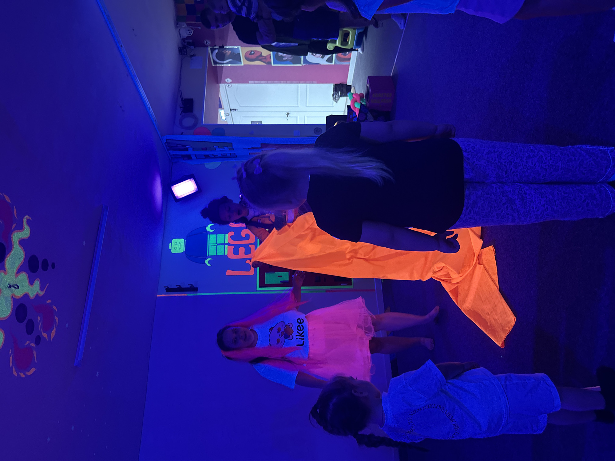Here is a description of the image:
**Scene:** An indoor space with blue and purple lighting, likely a room designed for blacklight or neon effects.
**People:** There are multiple children and at least one adult present. One child is wearing a white t-shirt with "Likee" written on it and a pink tutu, with pink hair. Another child is wearing a white top. An adult with blond hair is wearing a black top and patterned pants. An orange cloth is draped over some of the individuals.
**Objects:** Walls are decorated with fluorescent or glow-in-the-dark paintings and figures, including a "LEGO" design. There's a light source emitting a bright glow. The flooring is dark and the background includes posters and a door.
**Actions & Interactions:** The people appear to be interacting with each other, possibly during a party or event.
**Environment:** The room has a playful atmosphere due to the colorful decorations and lighting.
**Location:** The city is unknown.