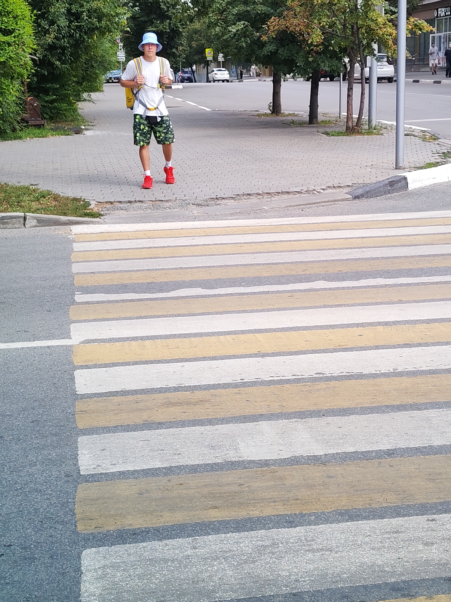 A young male wearing a blue bucket hat, a white t-shirt, and camouflage shorts walks across a pedestrian crossing. He has a yellow backpack on and is wearing red sneakers. The crossing is on an asphalt road with alternating yellow and white stripes. Trees and buildings are visible in the background, along with other pedestrians and vehicles. The scene appears to be on a street in a city.