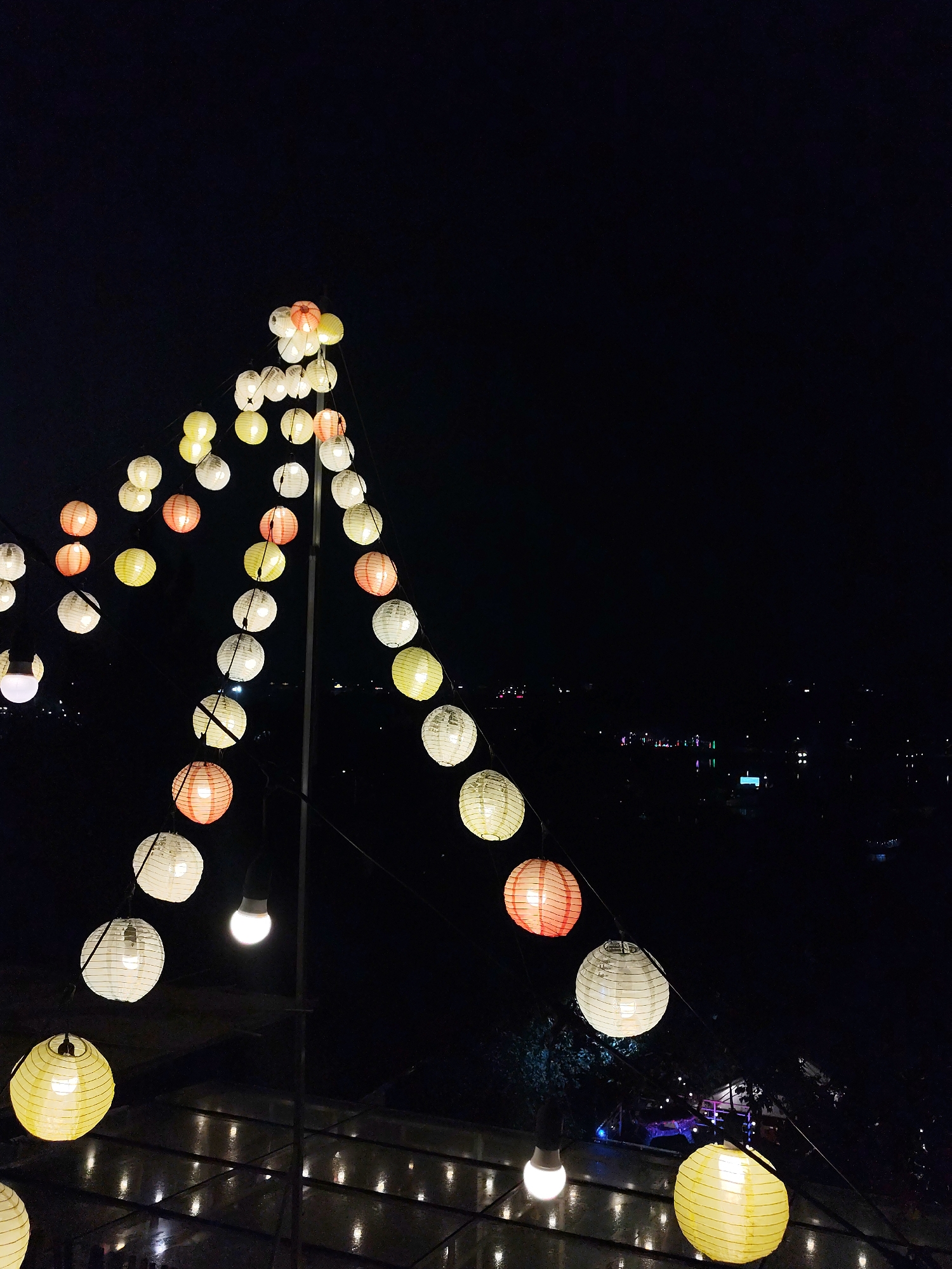At night, a low-angle medium shot captures strings of decorative lanterns. The strings form a tent-like structure in the foreground, with various colors including white, yellow, and red. Underneath, a grid-like translucent roof reflects the light from the lanterns and other light sources nearby. Beyond the lanterns, the dark outline of Da Lat is visible. Street lights are visible. The focus is primarily on the lanterns and roof.