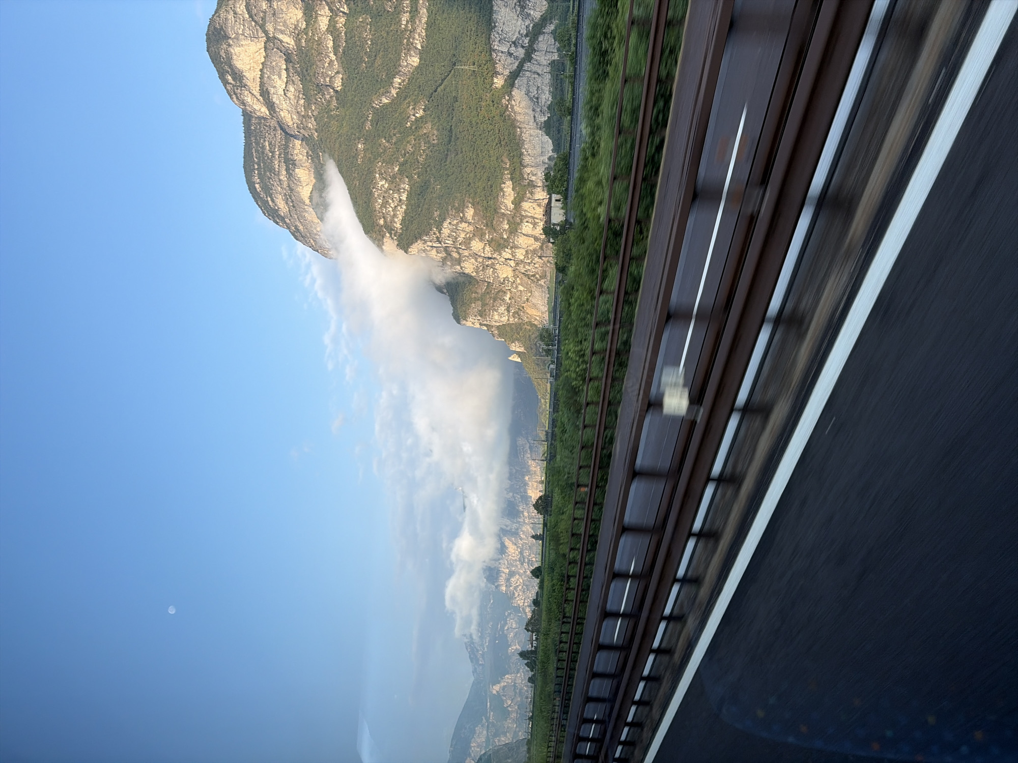 The image captures a mountainous landscape viewed from a highway. A highway with a white lane marking is in the foreground, with blurred motion suggesting it is being driven on. A rusty metal barrier separates the highway from a lush green field. Beyond the field, a rugged mountain range dominates the background, with the sun illuminating its rocky slopes and green vegetation. Clouds are clinging to the side of the mountain, obscuring parts of it. A small, pale moon is visible in the clear blue sky. In the distance, more mountains can be seen, hazy and receding into the background. A single building with a red roof is nestled at the base of the mountain. The scene appears to be in Neumarkt-Egna, Italy, given the characteristic Alpine scenery.