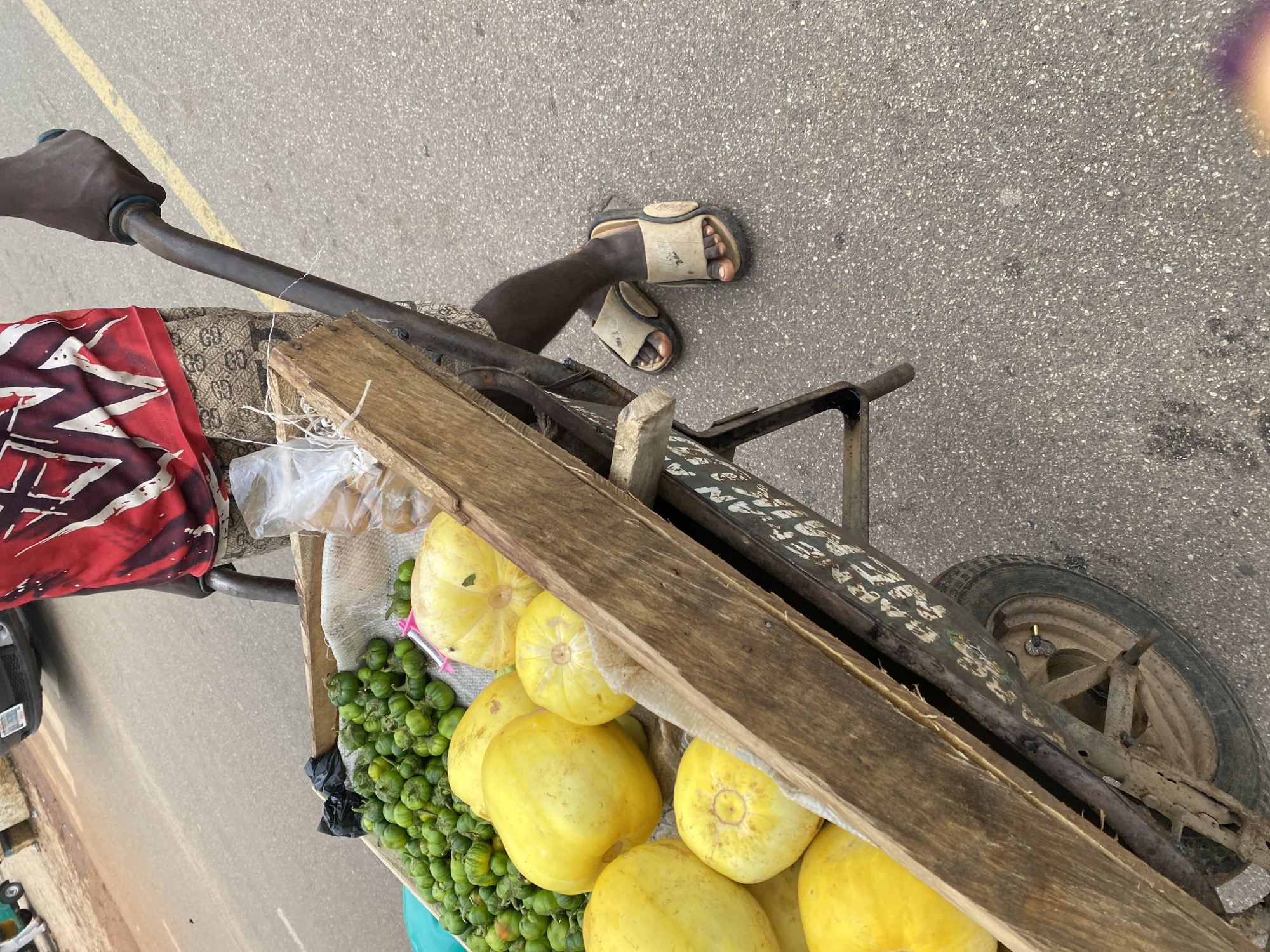 An outdoor, high-angle, medium shot captures a person pushing a wheelbarrow filled with fruits on a paved road in Rafin Par, Nigeria. The person, whose lower legs and feet are visible, wears patterned shorts and beige sandals. They are holding the handles of the wheelbarrow, which has a wooden frame and a metal structure. The wheelbarrow contains a variety of yellow fruits and a pile of small green vegetables. A plastic bag of what appears to be snacks is also present. The road surface is asphalt with painted yellow lines. In the background, a portion of a dark-colored car is visible. The overall lighting suggests it is daytime.