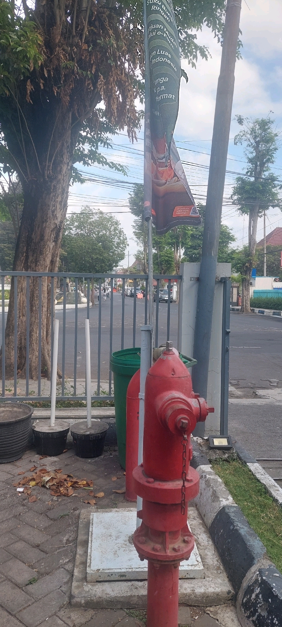 A red fire hydrant stands prominently on a paved sidewalk in Lumajang, Indonesia. The hydrant is positioned in front of a large gray metal fence, behind which are trees and a road with several cars and pedestrians. To the left of the hydrant are three black plant pots, two of which contain white poles cemented into them. A green trash can is situated behind the hydrant. A banner with Indonesian text and an image hangs from a pole to the right of the hydrant. The background shows more trees and buildings, with a blue sky overhead. The scene appears to be a street-level view of an urban environment.