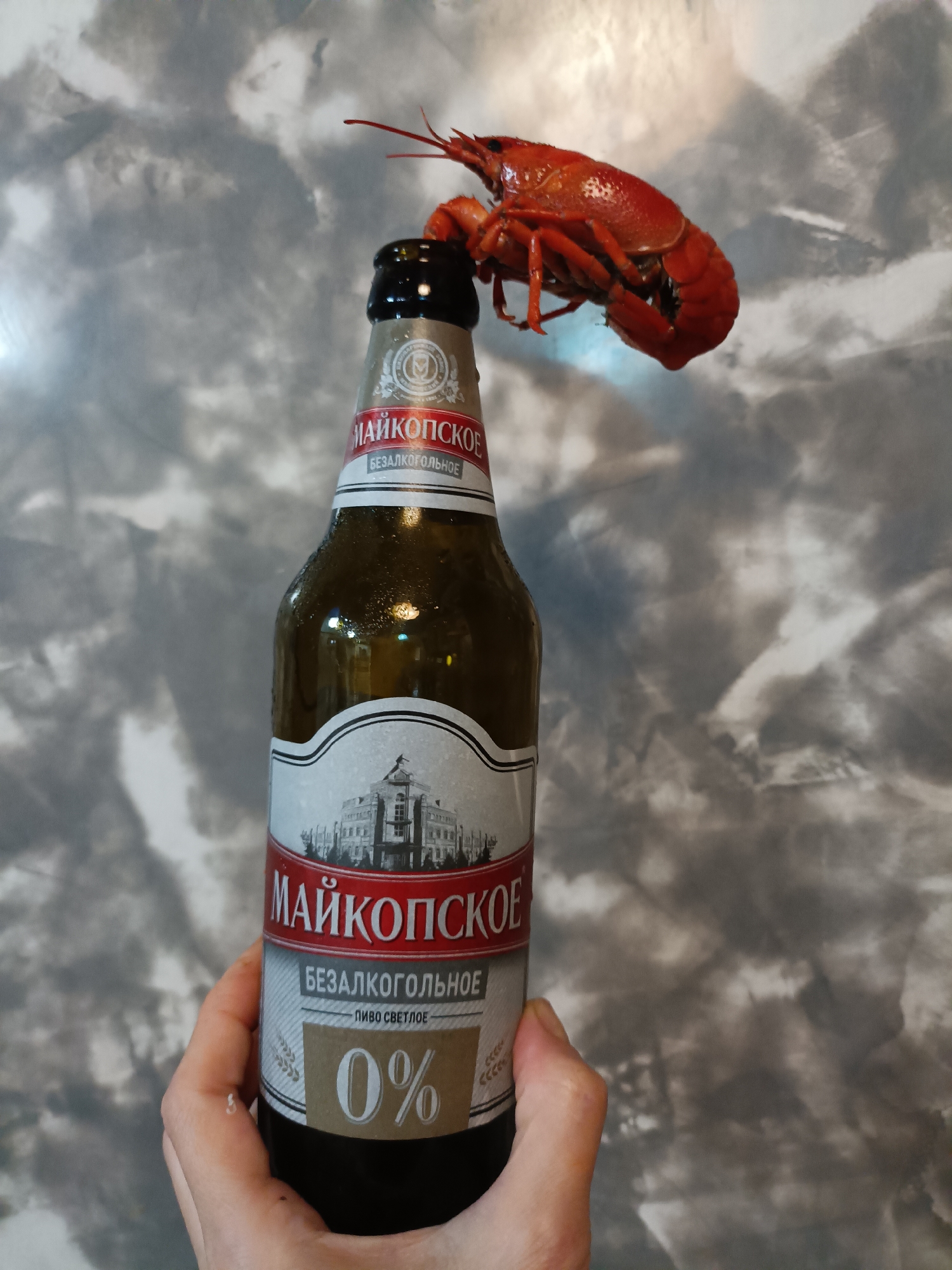 A crawfish is perched atop a bottle of "Maikopjskoe Bezalkogol'noe Pivo Svetloe" (Maykop Non-Alcoholic Light Beer) which has a 0% alcohol content. The bottle is being held by a person's left hand. The background is a textured, gray wall. The image was taken in Bataysk, Russia.