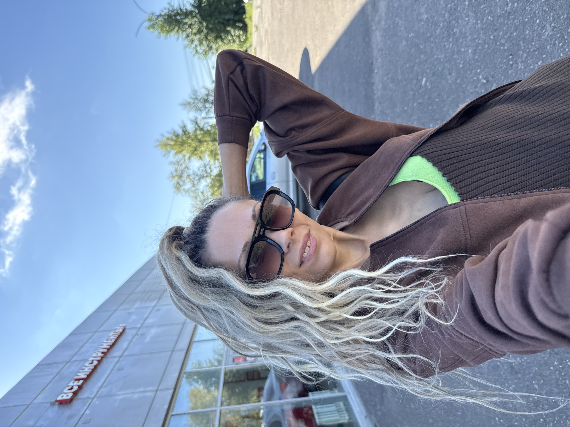 In Rusanovo, Russia, a woman with long, wavy blonde hair and sunglasses poses for a selfie outdoors. She is wearing a brown zip-up jacket over a neon green top and a brown ribbed dress. Behind her is a building with large windows and signage reading "ВСЕ ИНСТРУМЕНТЫ" in red letters, indicating a store. A car is visible through the windows. The scene is set against a clear blue sky with scattered clouds, and some trees are visible in the background.