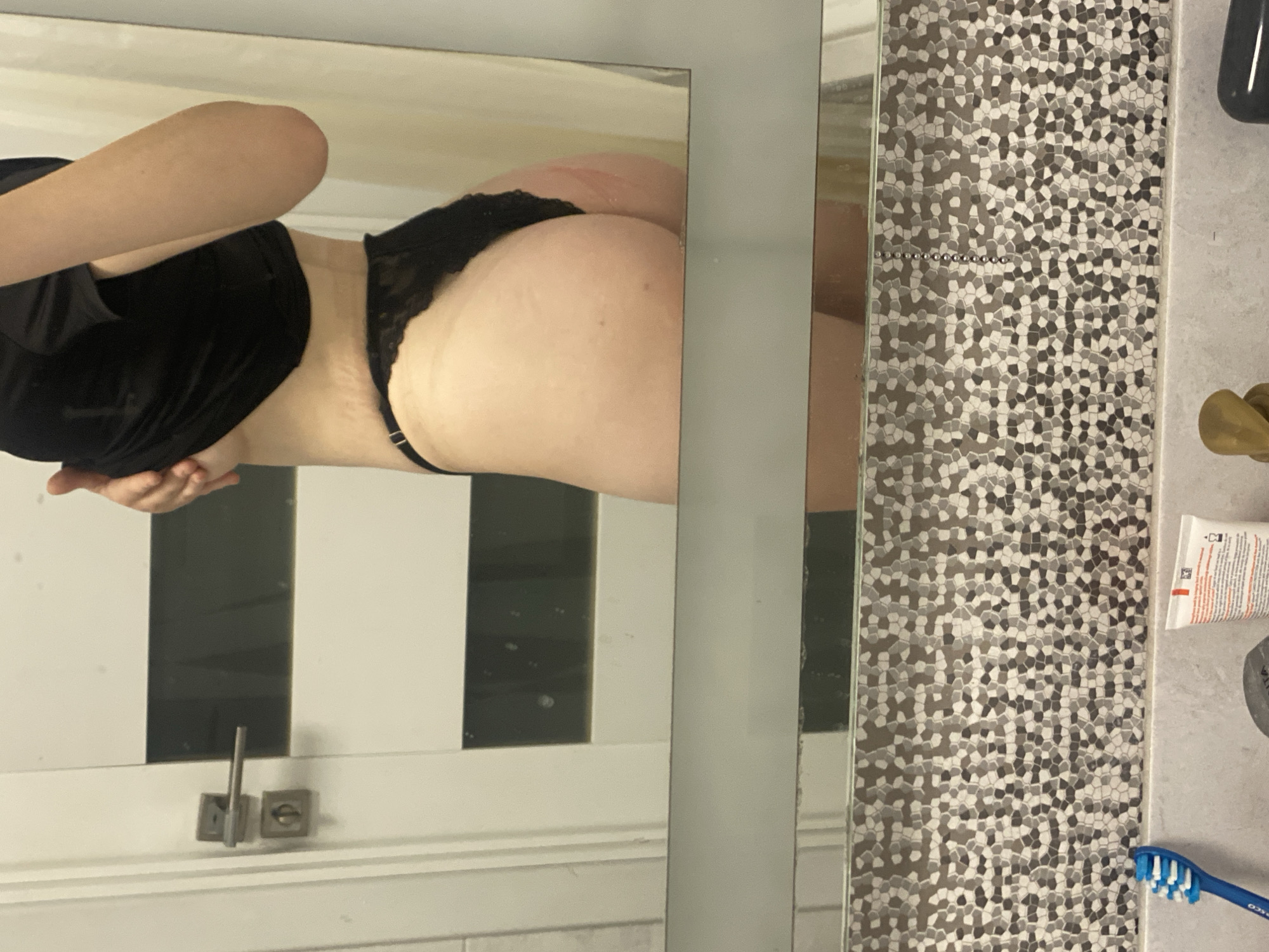 The image captures a rotated view of a bathroom scene, with the main subject being a person reflected in a large mirror. The person, from approximately the mid-torso downwards, is wearing a black, possibly velvet, short-sleeved top and black lace thong-style underwear. Their left arm is bent, with their hand partially visible at the bottom of the frame, suggesting they are holding a device to take a mirror selfie.
The mirror on the left reflects what appears to be a white door or cabinet with dark vertical panels behind the person. At the bottom of this reflected section, there are two square metal fixtures, possibly door handles or locks. The mirror itself has a thin grey frame on the left and a wider, solid grey frame separating it from a section of wall. This wall is adorned with intricate mosaic tiles, consisting of small, irregularly shaped pieces in various shades of grey, black, and white.
To the right of the tiled wall, a countertop is visible, holding several bathroom essentials. These include a blue toothbrush with white bristles, on which the text "OSCO" (possibly part of a brand name like "CURAPROX" or similar) is partially legible. Next to it is a white tube of toothpaste, showing the partial text "COLGAT" (likely "COLGATE") and a brand logo with "N." and other small text. A golden or bronze-colored object, possibly a tap or a decorative item, and a dark cylindrical container are also on the counter. The overall setting is a modern bathroom in Ternopil, Ukraine.