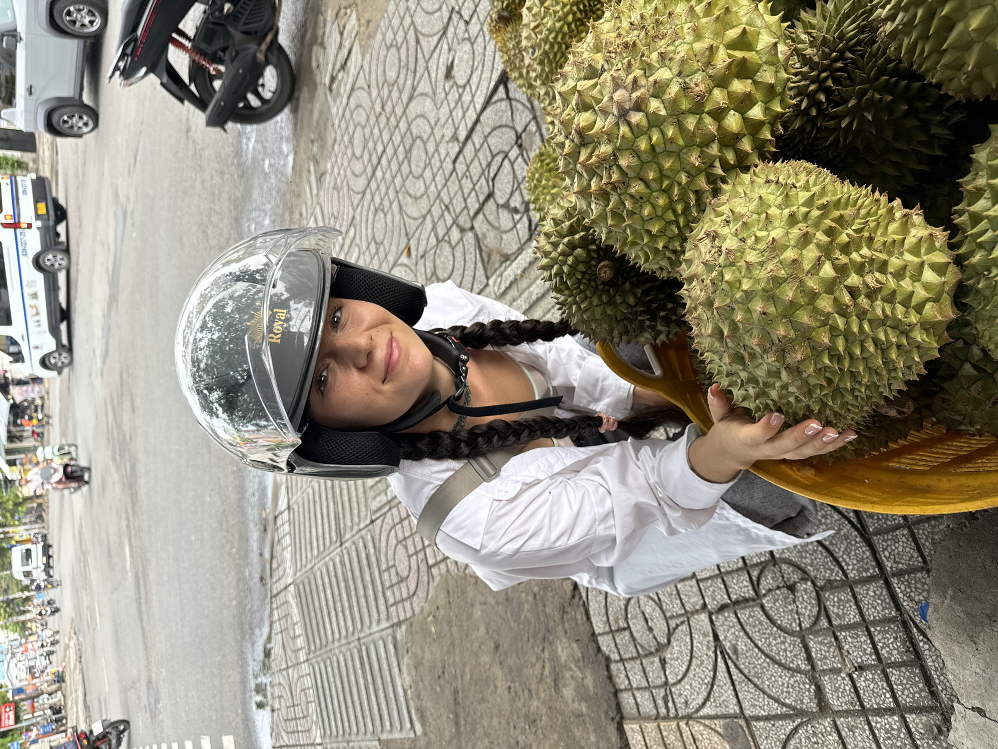 A young woman with long dark braids, wearing a clear motorcycle helmet and a white shirt, is standing next to a basket overflowing with spiky durian fruits in Phu Thuy Ward, Vietnam. She has one hand resting on a durian. The background includes a street with a motorcycle, vehicles, and a sidewalk with patterned tiles.