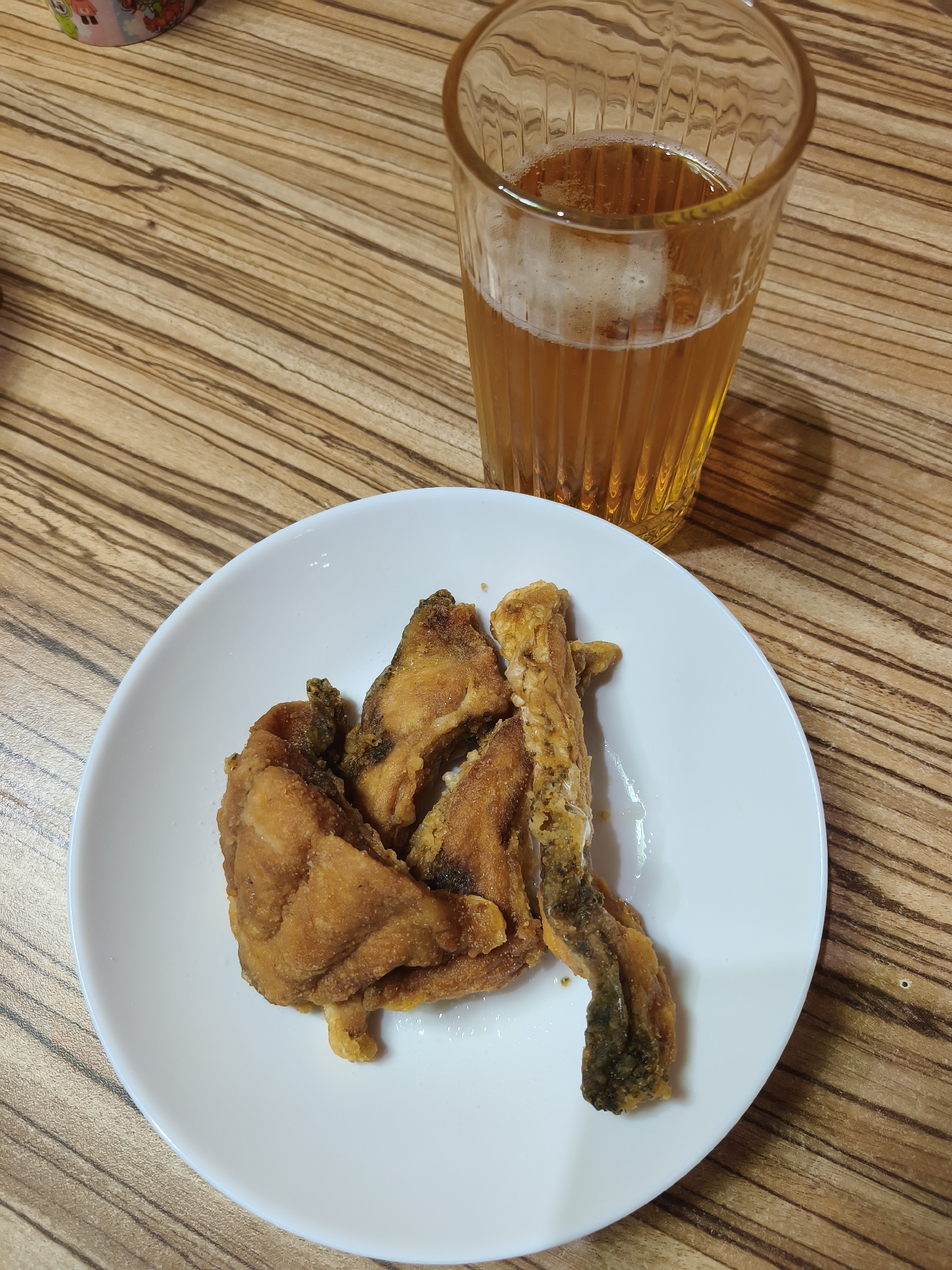 A close-up, elevated shot captures a casual meal set on a wooden table. A white, round plate prominently displays several pieces of golden-brown fried fish, suggesting a crispy texture. One elongated piece on the right side of the plate clearly shows a darker, possibly skin, portion. Adjacent to the plate, in the upper right quadrant of the image, stands a clear glass filled with a golden-amber beverage, topped with a frothy white head, characteristic of beer, ale, or lager. The glass has a distinct ribbed or fluted design. The table's surface features a prominent, light brown wooden grain with darker, wavy patterns. The lighting suggests an indoor setting, possibly during the day or evening. No people are visible, and there is no discernible text. The scene depicts a simple, prepared meal or snack, set in what is identified as Svilengrad, Bulgaria.