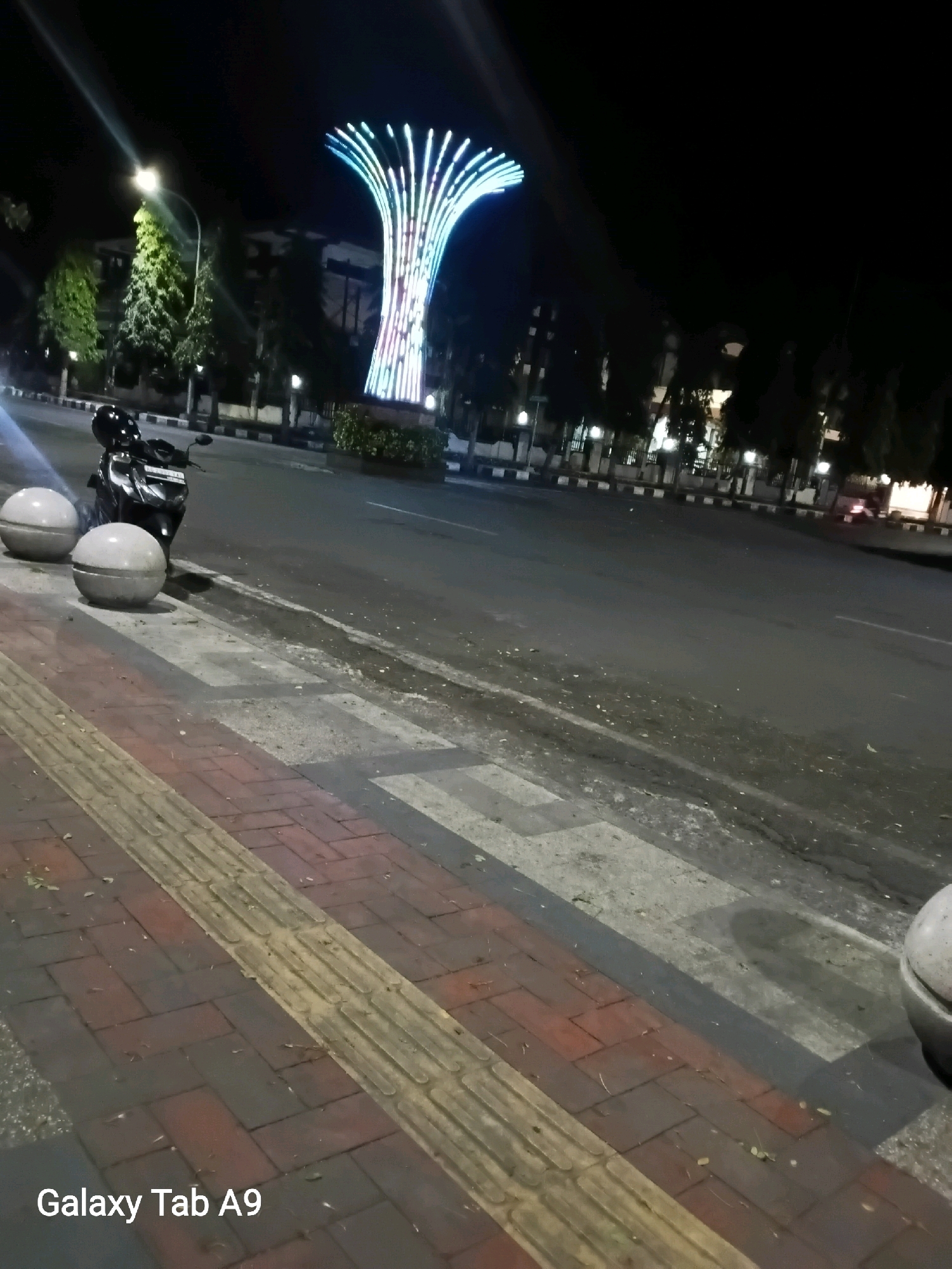 A night time exterior shot captures a street scene in Trenggalek, Indonesia. In the background, a large, tree-like sculpture is illuminated with colorful lights, featuring vertical columns of blue, green, and pink. The sculpture is situated behind a row of trees and buildings, some of which are also lit. In the foreground, a motorcycle with a black helmet on its seat is parked on the side of the road. Two large, spherical concrete bollards are positioned next to the motorcycle. The sidewalk in the immediate foreground is paved with red bricks and features a tactile paving strip. The road appears wet or damp, reflecting the surrounding lights. The overall atmosphere is calm and urban. The text "Galaxy Tab A9" is visible in the lower left corner of the image.