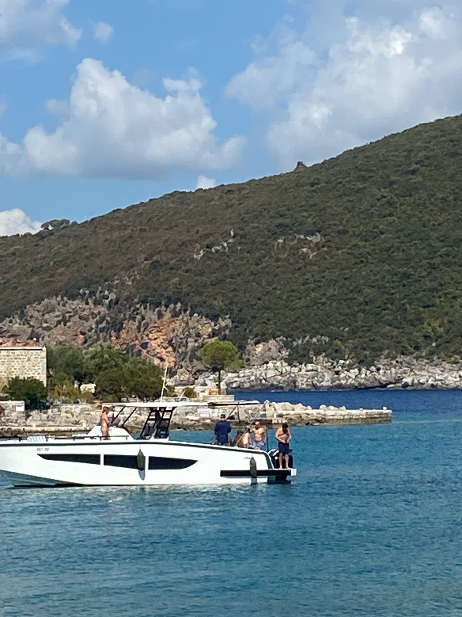 A white speed boat with black accents is positioned in the turquoise waters of Luštica, Montenegro. Several individuals are visible on the boat; one person stands near the bow, another sits on a bench, and two individuals are at the stern, one appearing to be taking a picture. The boat is in the foreground, with a rugged, tree-covered hillside rising behind it. The hillside is characterized by rocky outcrops and dense vegetation. Along the shoreline, there is a stone building and a breakwater made of large stones. The sky above is a clear blue with scattered white clouds.