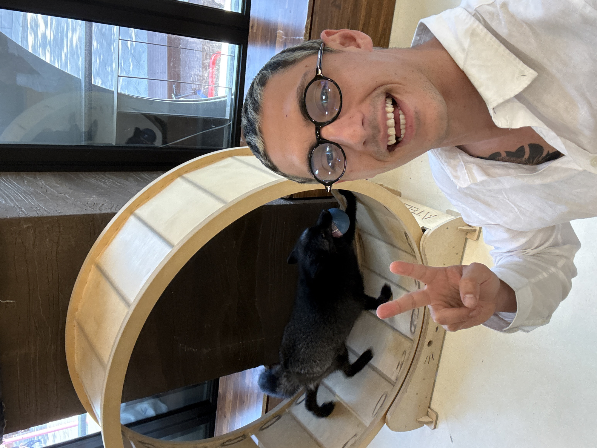 The image shows a man taking a selfie with a black fox in a cat wheel. The man is wearing glasses and a white shirt, and making a peace sign. He has a tattoo on his chest. The fox is playing with a blue ball inside the wheel. The background includes a window and part of a wooden wall. The photo appears to be taken indoors. The precise location in Seoul, South Korea, cannot be determined from the image.