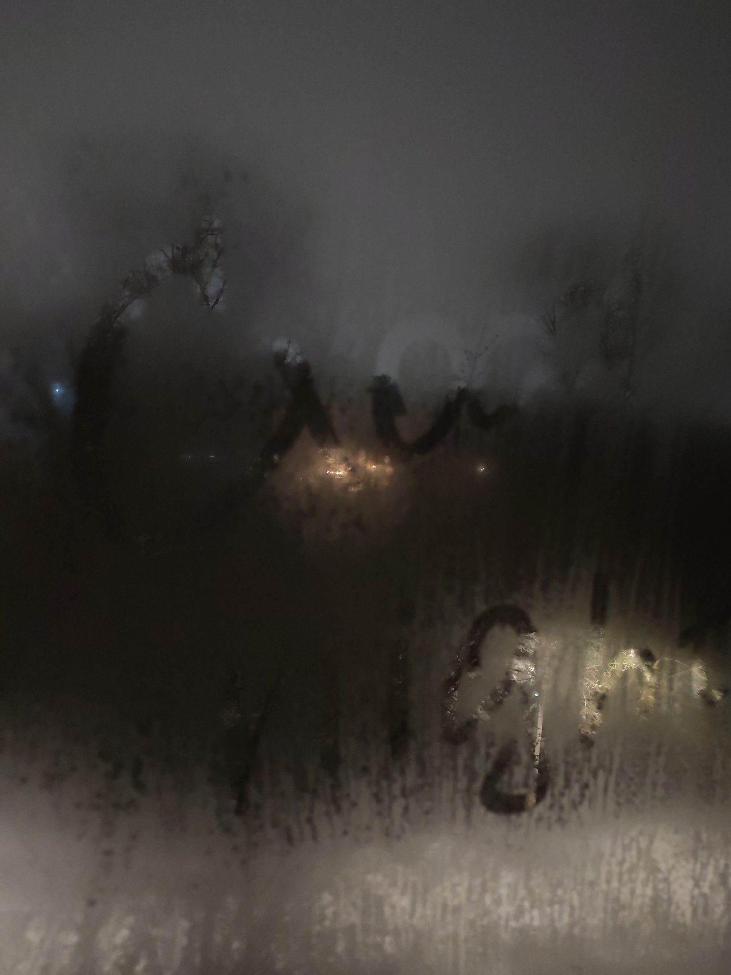 The image depicts a view through a fogged-up window, likely on a cold or humid night. Writing, perhaps "Good Night", is partially visible on the glass. Outside, blurred lights suggest a city street scene. Judging from the prompt, the location may be Izhevsk, Russia, though this is not visually verifiable. No people are visible, and details of specific objects outside are obscured by the condensation on the window. The primary subject is the interaction between indoor condensation and outdoor light, creating a blurred, atmospheric scene.