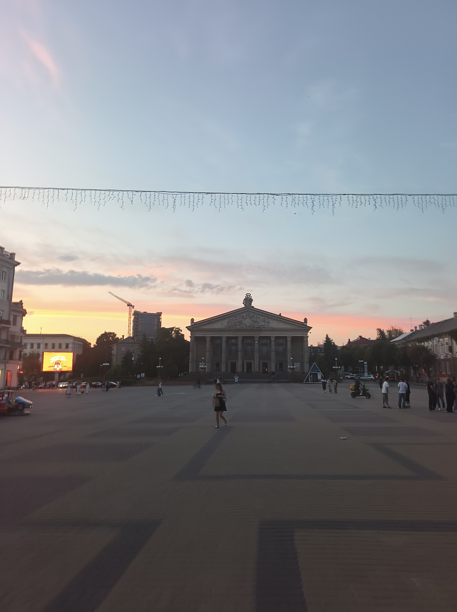 The image depicts Teatralna Square in Ternopil, Ukraine, during the early evening. The sky displays soft pastel hues of pink and orange from a sunset, with a clear blue gradient extending upwards. A string of decorative lights hangs across the upper portion of the frame.
Dominating the center of the image is the Ternopil Regional Drama Theater, a grand building with a classical facade featuring a portico and columns. A statue is visible atop its roof. To the left, a large outdoor LED screen displays vibrant graphics. Further left, a multi-story building with visible windows lines the square. A construction crane and a partially built structure are visible in the middle distance, beyond the screen and to the left of the theater.
The square itself is paved with patterned paving stones, with darker rectangular sections forming a visual design. Several people are present in the square. In the foreground, a woman in a dark dress walks towards the right. Further back, a group of people are gathered near the theater entrance. A yellow scooter is parked in the middle ground, and several other individuals and small groups are scattered across the open space. Cars and a small amusement ride are visible on the left side of the square. The overall atmosphere suggests a relaxed evening in a public urban space.