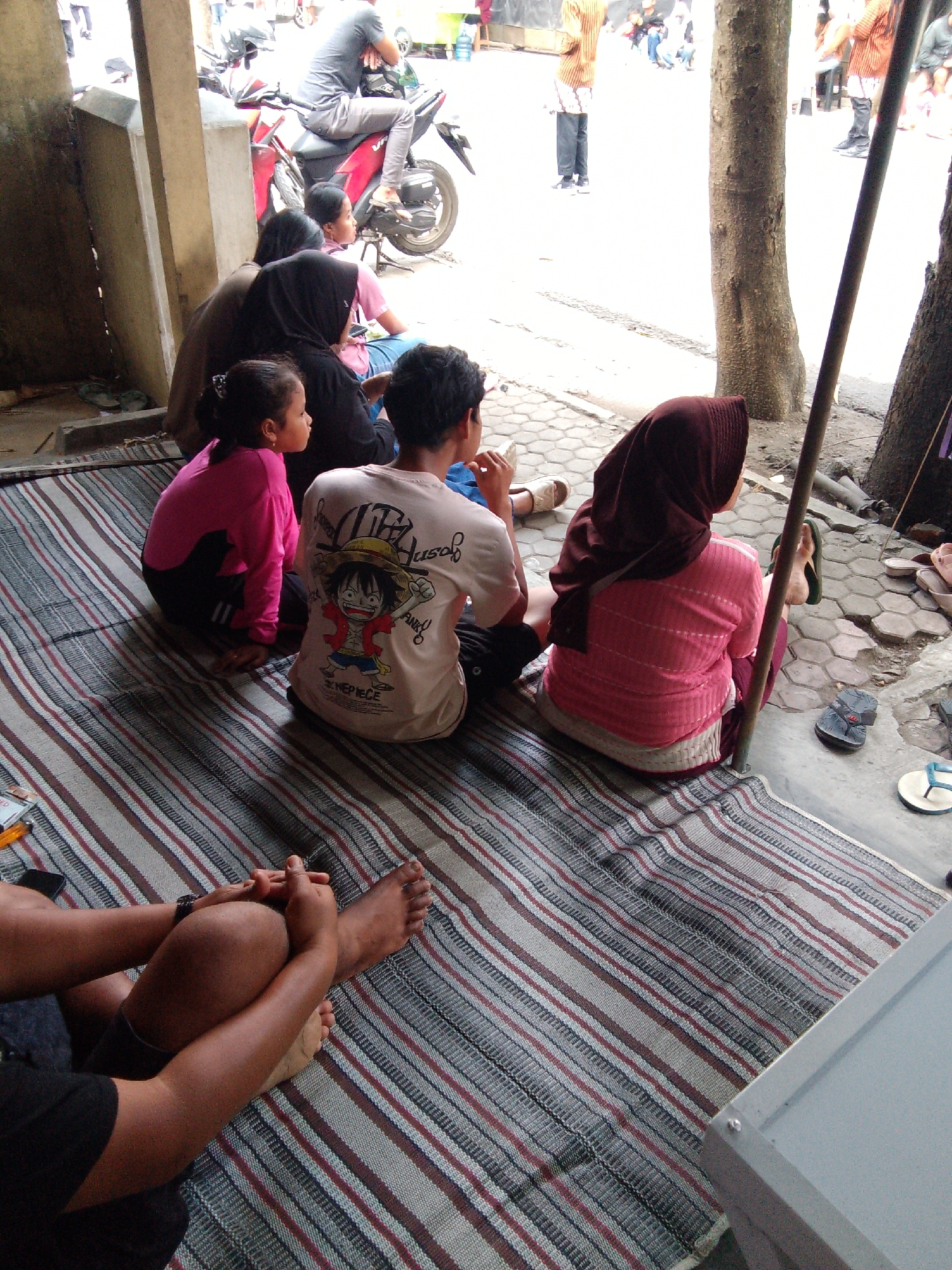 The image depicts a group of people sitting on a striped mat on the ground in what appears to be a public area, possibly a street or a market stall in Trenggalek, Indonesia. In the background, a red motorcycle is visible with a person sitting on it. Several other individuals are standing and walking in the background, some wearing traditional Indonesian clothing. The foreground shows four people sitting on the mat: a young girl in a pink long-sleeved shirt and black pants, a young man wearing a light-colored t-shirt with an anime character graphic, a woman wearing a brown headscarf and a pink long-sleeved shirt, and another person partially visible at the bottom of the frame. The environment suggests a warm climate, with some trees visible in the background and paved ground. The overall scene conveys a sense of everyday life and community gathering.