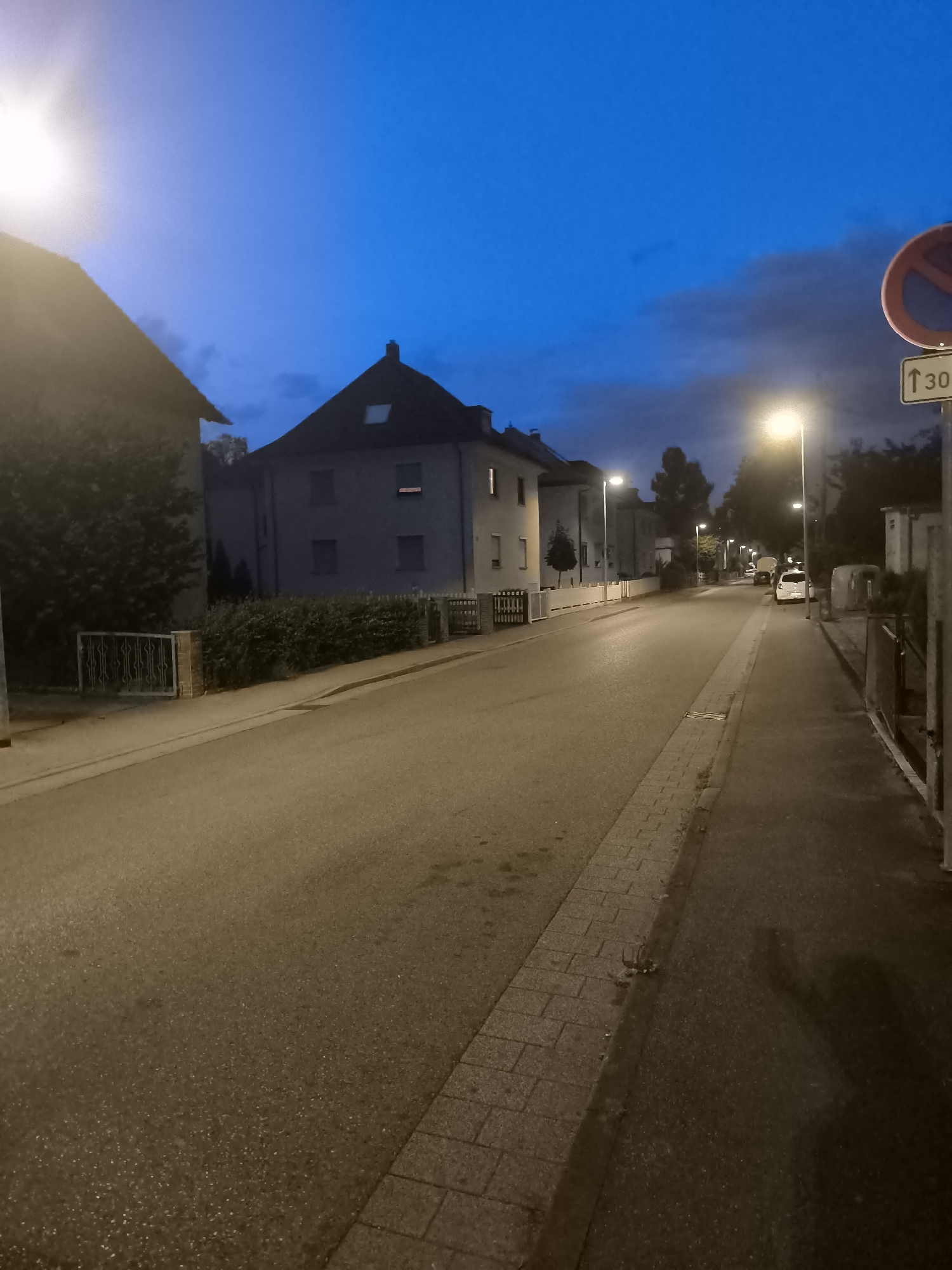 An outdoor, eye-level, vertical medium shot shows a paved street at dusk in a residential area. The street is lit by streetlights, and a bright light source is visible in the upper left corner. The sky is a deep blue with some clouds. On the left side of the street, there is a building with a dark roof and several windows. A fence and bushes are in front of the building. On the right side of the street, there is a sidewalk with a curb, and a few parked cars are visible further down the street. A traffic sign is also present on the right side of the street. The pavement appears wet in some areas.