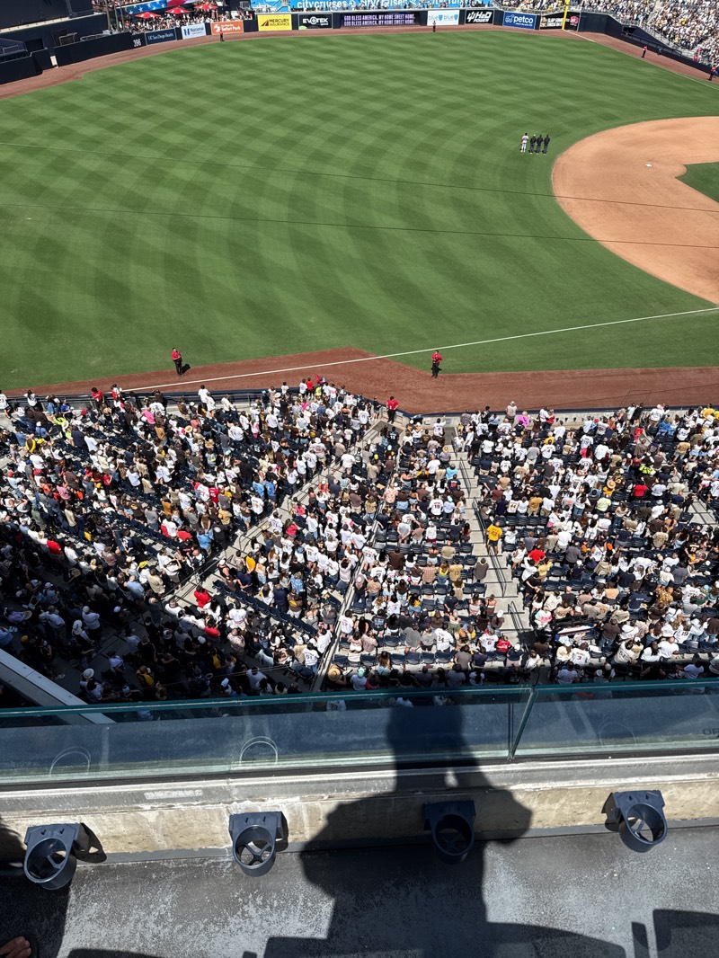 The image depicts a baseball game in progress, viewed from an elevated position overlooking the field and a large crowd of spectators. The field is well-maintained, with neatly striped grass and a brown infield. A small group of people are standing on the infield near the first base line. The crowd fills the stands in the foreground, densely packed and varied in attire. A glass barrier is visible in the bottom foreground, with cup holders attached to the concrete wall. The shadow of a person or object is cast onto the concrete surface in the bottom center. The stadium's location is in San Diego, United States, as evidenced by the "San Diego" signage on the outfield wall. The weather appears sunny and bright, suggesting a daytime game.