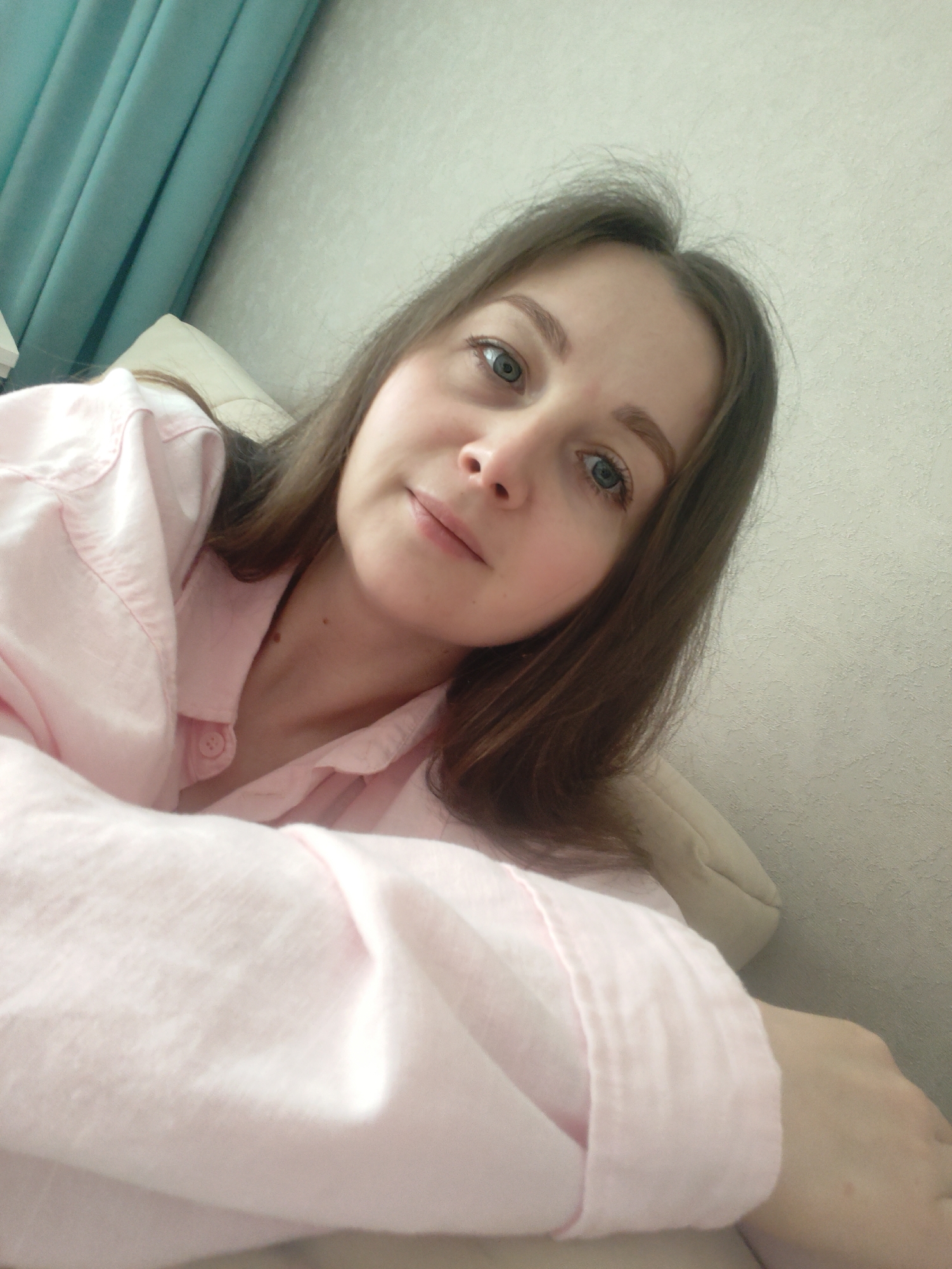 A young woman is featured in a close-up, slightly high-angle selfie. She has long, dark brown hair, parted subtly off-center, framing her face. Her eyes are a light blue or grey, and she has a calm, relaxed expression with a faint, closed-lip smile. She is wearing a light pink, collared, long-sleeved shirt, possibly made of a soft, cotton-like material, visible in the foreground and around her upper body.
The background shows an indoor setting. To the left, there's a soft teal or light blue curtain, suggesting a window nearby. The rest of the background consists of a light-colored wall with a subtle, textured pattern. A light-colored pillow or cushion is partially visible behind her head and shoulder, indicating she might be lounging on a bed or sofa. Her right arm, covered by the pink sleeve, is prominent in the lower left portion of the frame. The overall lighting is soft, creating a gentle and casual atmosphere. There is no visible text in the image.