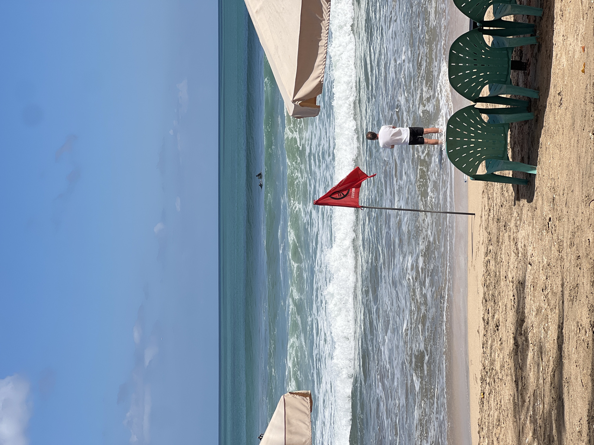 In Kuta, Indonesia, on a bright day, a man in a white t-shirt and black shorts stands in the shallow ocean, facing away from the camera. A red flag with the word "SWIM" is planted in the sand in front of him. Two surfboards and two surfers are visible in the distance, on the waves. Two beach umbrellas, one on the left and one on the right, provide shade on the sandy beach, where several green plastic chairs are arranged. The ocean is turquoise with white-capped waves. The sky is blue with a few scattered white clouds.