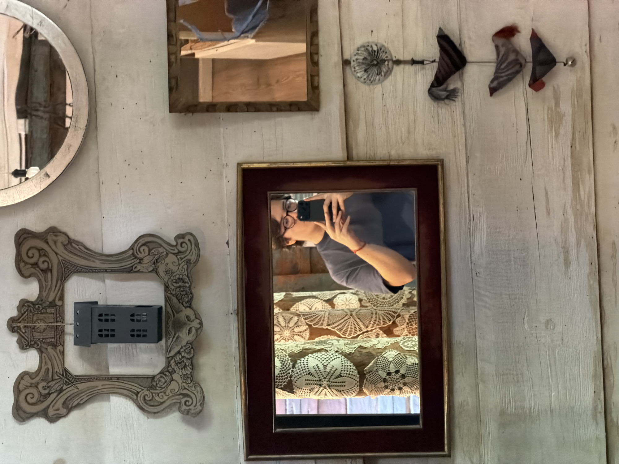 The image captures an indoor scene, likely in a shop or home in Puebla, Mexico, adorned with various mirrors and decorative frames on a light-colored, distressed wooden plank wall.
The central subject is a person, reflected in a rectangular mirror with a dark red and gold frame, holding a black smartphone to take the photograph. The person wears glasses and a light blue/grey shirt, with a red band or bracelet visible on their wrist. Below the person's reflection, the mirror reveals a stack of numerous white, intricately crocheted doilies, suggesting a display or collection.
To the upper left, an oval mirror with a silver-colored, textured frame reflects indistinct shapes, possibly clothing or parts of the room. Above the central mirror, a smaller rectangular mirror with a brown wooden frame reflects what appears to be wooden shelving or furniture.
On the lower left, an ornate, empty frame with an elaborate, possibly baroque or Art Nouveau design, holds a black, rectangular object resembling a miniature building or an electronic device with small, gridded windows. To the upper right, a decorative piece features three fabric "fish" or arrow-like shapes, made of patterned and striped cloth, strung together by a wire extending from a central, round, ornamental element.
The overall ambiance is one of curated vintage or rustic decor. There is no visible text in the image.
