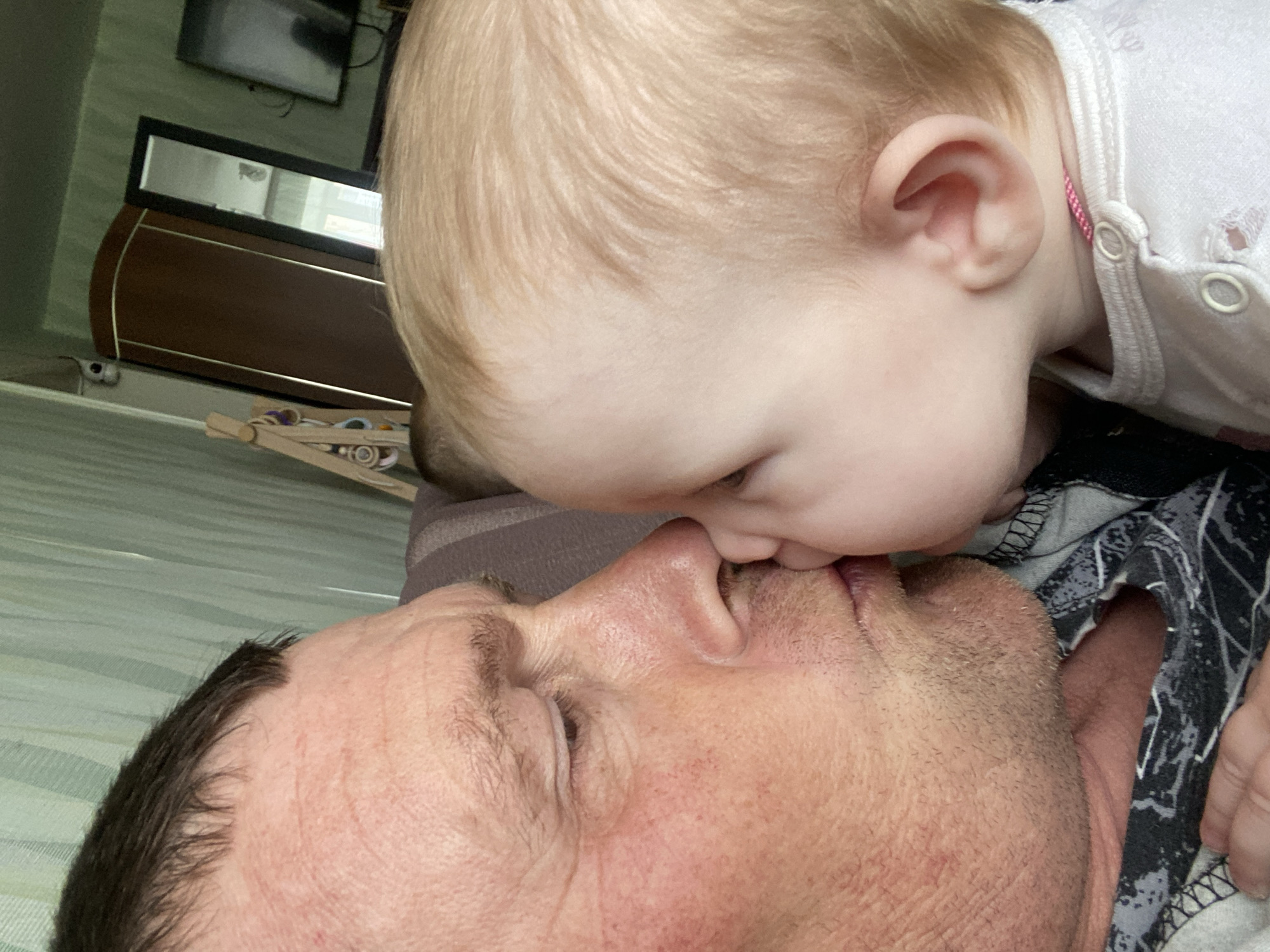 A close-up shot shows a man lying on his back with a baby leaning over him, their faces touching. The baby has blonde hair and is wearing a white top with buttons. The man has dark hair, and the image is taken at a close angle, focusing on the facial interaction between the two. The background includes a dresser with a mirror and a television on the wall, suggesting an indoor setting. The location cannot be determined from the image alone.