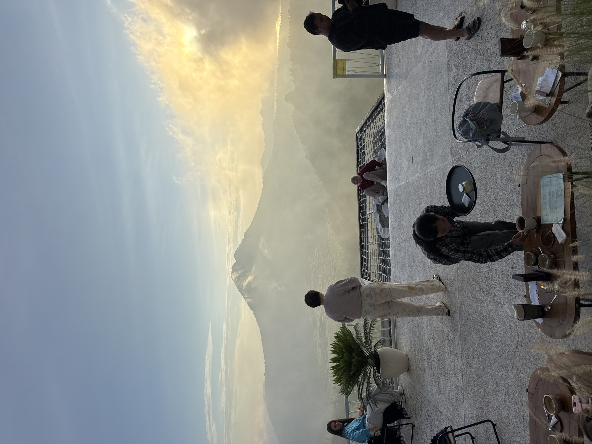 The scene is set in a cafe in Kedisan, Indonesia, with a clear view of Mount Batur on a partly cloudy day. The lighting suggests it might be sunrise or sunset. The sky graduates from blue to golden yellow near the horizon where the sun might be hidden. People are scattered on a terrace. One man standing on the right of the frame is taking a photograph with a camera. Next to him are tables and chairs where people are sitting. Other people stand and look at the mountain. In the foreground, there is a palm tree in a large white pot. The overall atmosphere is calm and relaxed, as people enjoy the view and each other's company in the cafe setting.
