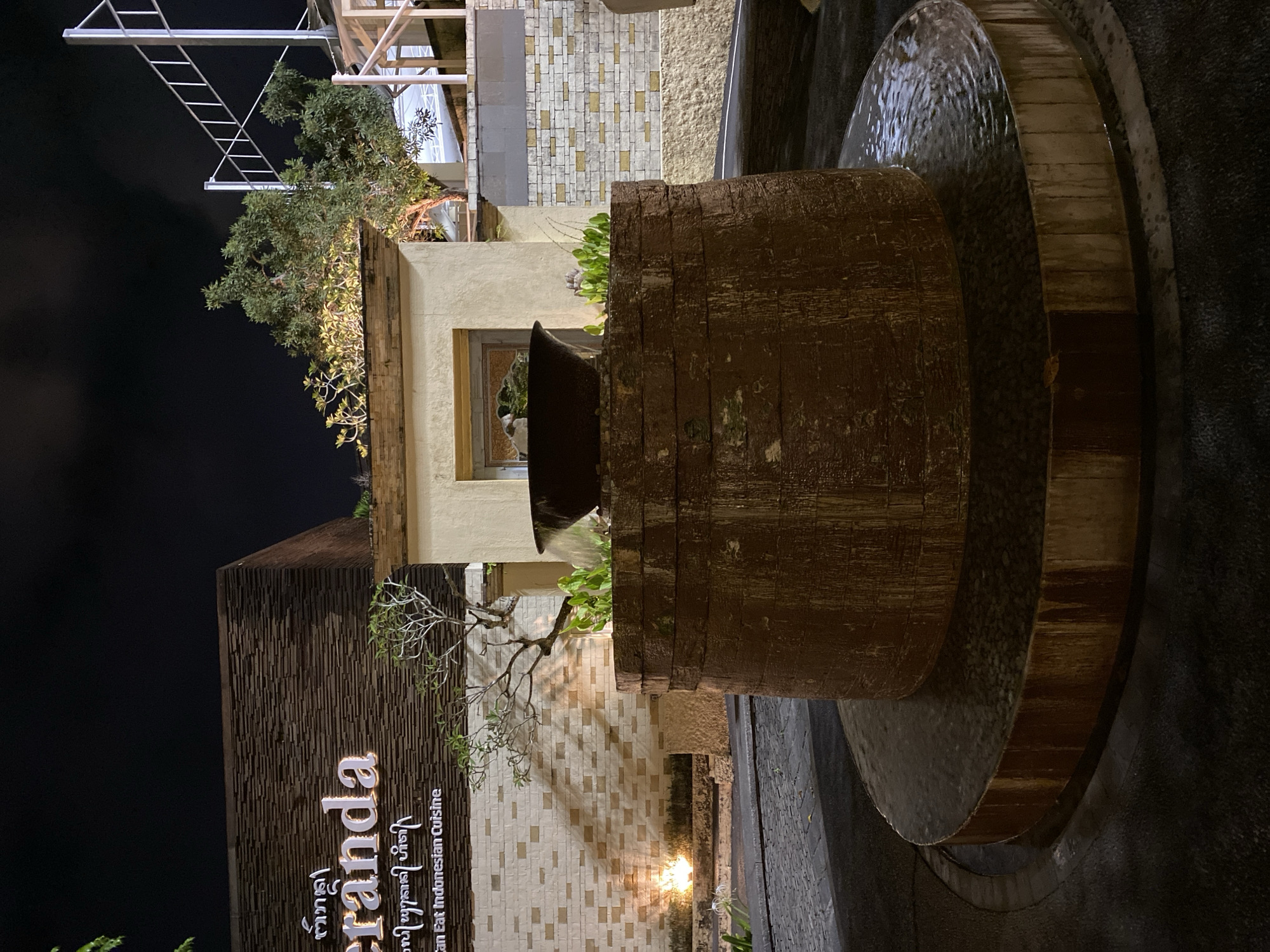 The image depicts the exterior of a restaurant at night, with a focus on a tiered water fountain in the foreground. The restaurant's sign, "Teranda Can Eat Indonesian Cuisine," is visible on a textured wall. The fountain is made of dark, possibly stone or wood, material and is surrounded by dark pavement and cobblestones. Plants are visible around the fountain and on top of the restaurant's structure. The environment is lit by artificial lights, casting shadows on the brick wall behind the fountain. The scene appears to be in a tropical setting, consistent with Kuta Selatan, Indonesia, known for its dining establishments and vibrant nightlife.