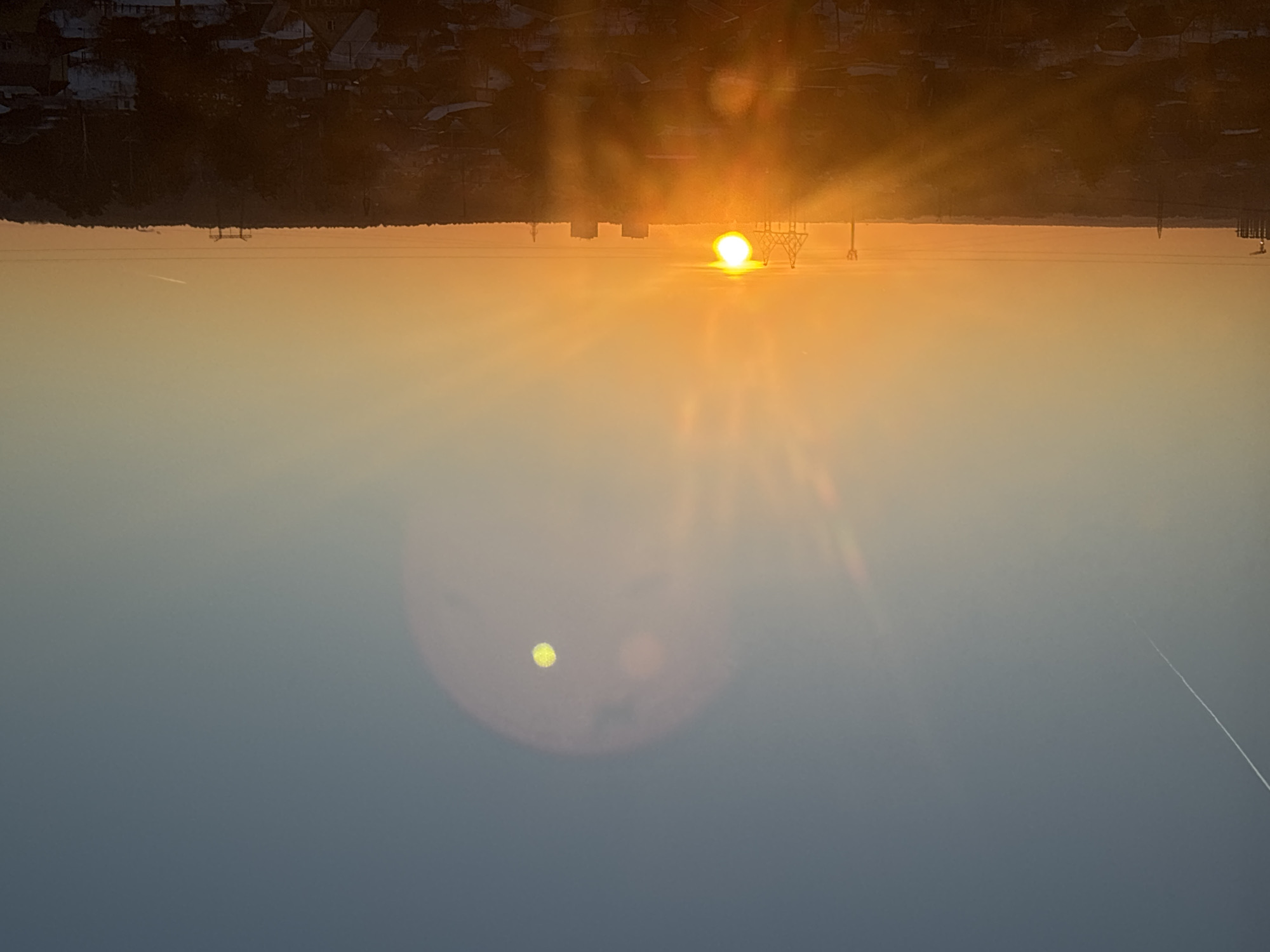 The image displays an elevated view of the city of Chaikovsky, Russia, during either sunrise or sunset. The sky exhibits a gradient from dark blue at the top to a vivid orange at the horizon, where the sun, appearing as a brilliant yellow-orange orb, is positioned slightly left of center. Intense solar glare and lens flares emanate from the sun, projecting bright light downwards. A distinct, bright yellow, circular optical artifact with a textured surface is visible in the upper-center sky, enclosed within a larger, diffuse reddish-purple lens flare. A thin, white contrail diagonally traverses the sky from the upper-left. The horizon line consists of a dark silhouette of industrial structures, including multiple power transmission towers, several rectangular multi-story buildings, and a dense treeline. Horizontal power lines are visible across the midground. The foreground is composed of dark, silhouetted rooftops of numerous residential buildings, interspersed with patches of white snow, indicating a cold season.