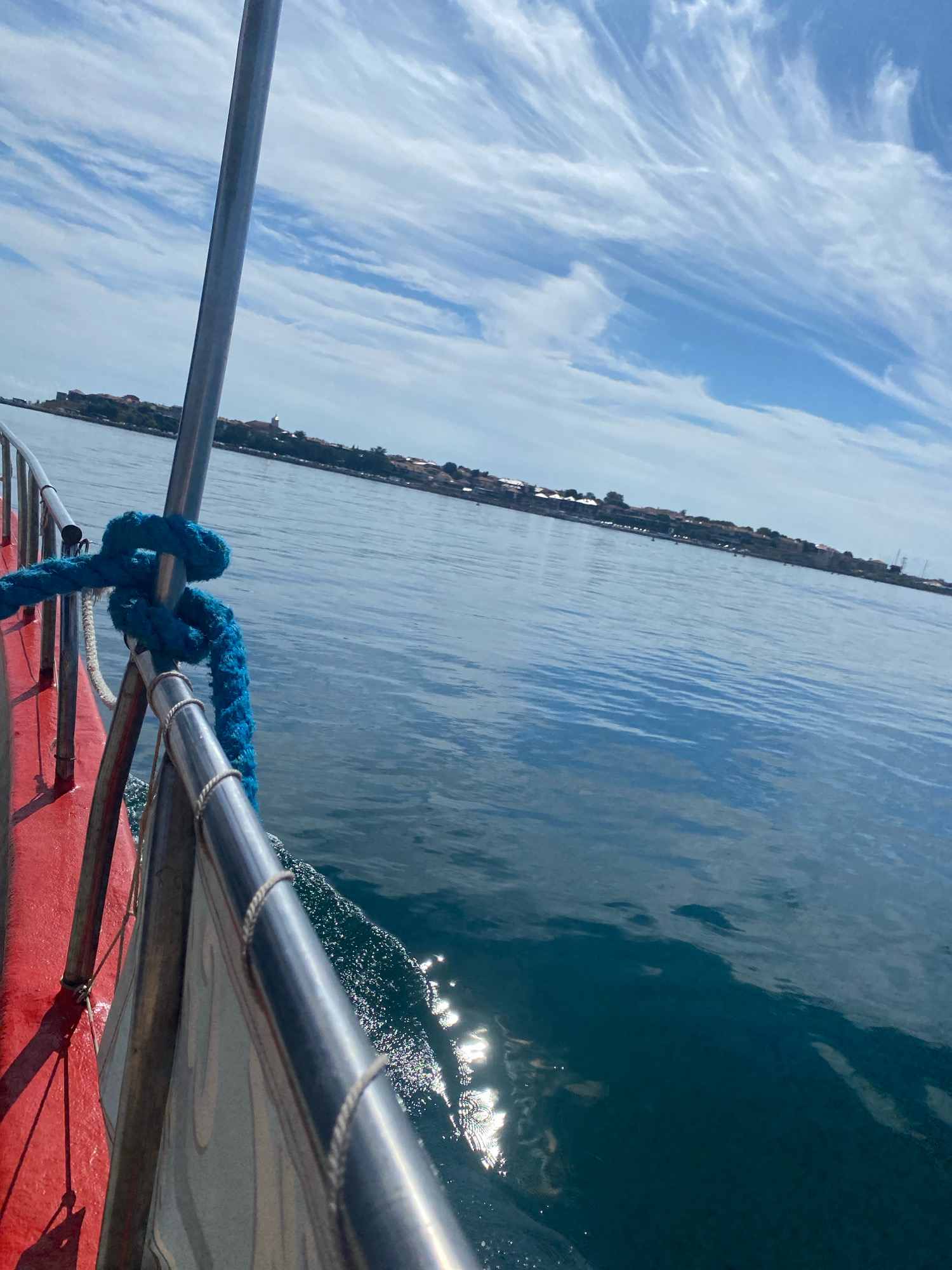 This image shows a view from a boat traveling on the sea towards Nesebar, Bulgaria. The boat's railing, painted red and adorned with a blue rope, is in the foreground. The water is a deep blue with sunlight glinting off its surface. In the distance, the historic town of Nesebar can be seen, with its distinctive red-roofed buildings nestled along the coastline under a bright blue sky with wispy white clouds. The overall scene depicts a calm day at sea, approaching a picturesque coastal town.
