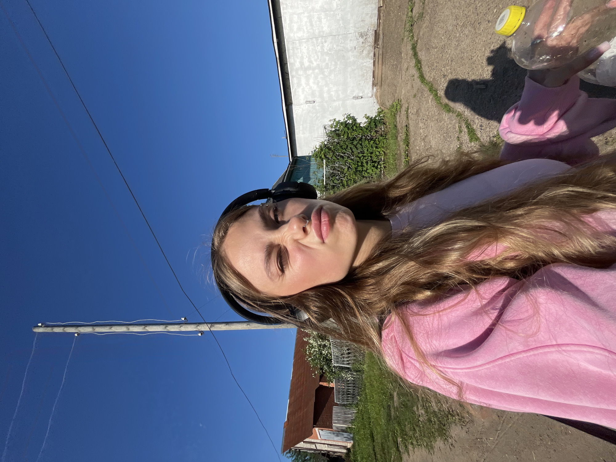 A young woman with long blonde hair and headphones on her ears is taking a selfie outdoors. She is wearing a pink hoodie over a light pink shirt. Her facial expression is a pout. She is holding a partially filled plastic water bottle with a yellow cap in her right hand. The background shows a rural setting with a dirt road, a wooden fence, a red-roofed building, a white garage-like structure, and a utility pole with wires. The sky is clear and blue. The image appears to be taken in the city of Fokino, Russia, given the visual context of the rural structures and utility pole, consistent with the typical appearance of such infrastructure in that region.