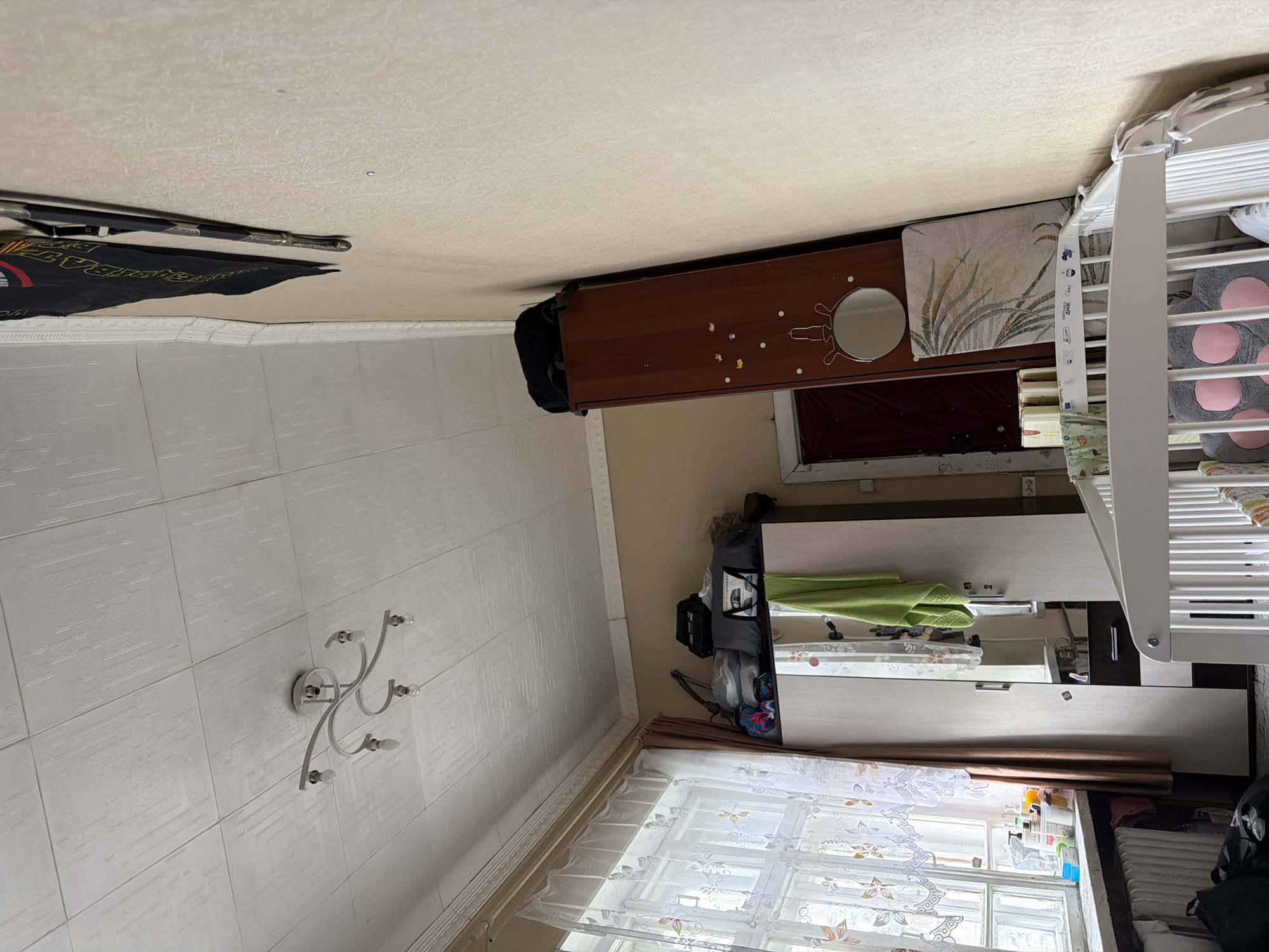 The image shows an interior scene, likely a bedroom, with a focus on furniture and decor. The room has a textured white ceiling with a modern, multi-arm light fixture. The walls are a neutral beige color. To the left, a large window with sheer, patterned curtains and a brown curtain allows natural light into the room. Below the window is a white radiator and some bags on the floor. In the center of the image is a tall, light-colored wardrobe with mirrored doors. A green towel is draped over the wardrobe. On top of the wardrobe, several items are stored, including bags and what appears to be a lamp. To the right of the wardrobe, a tall, narrow, dark wood cabinet leans against the wall. A round mirror is affixed to this cabinet, and a framed artwork is placed in front of it. A portion of a white crib is visible in the foreground, with bedding and a stuffed animal. The overall impression is of a functional, lived-in space, possibly a child's room, with a somewhat cluttered but organized arrangement of items. The scene does not contain any specific indicators of being located in Moscow, Russia.