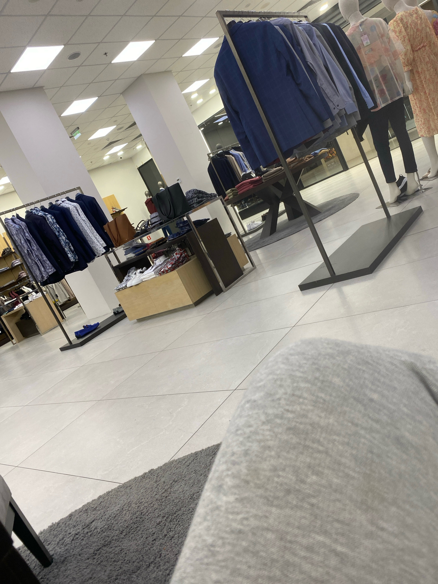 An indoor shot captures a retail store, likely a clothing boutique, in an unknown city. The scene is dominated by clothing racks filled with menswear, primarily suits and shirts in various shades of blue and white. Mannequins dressed in female attire, including a light yellow patterned dress and a sheer, long-sleeved garment, stand to the right. The store features clean, tiled flooring, illuminated by overhead fluorescent lighting. In the foreground, a person's grey sweatpants and a section of a grey rug are visible, suggesting the viewpoint of someone seated within the store. Other elements include display shelves with folded clothing, handbags, and shoes.