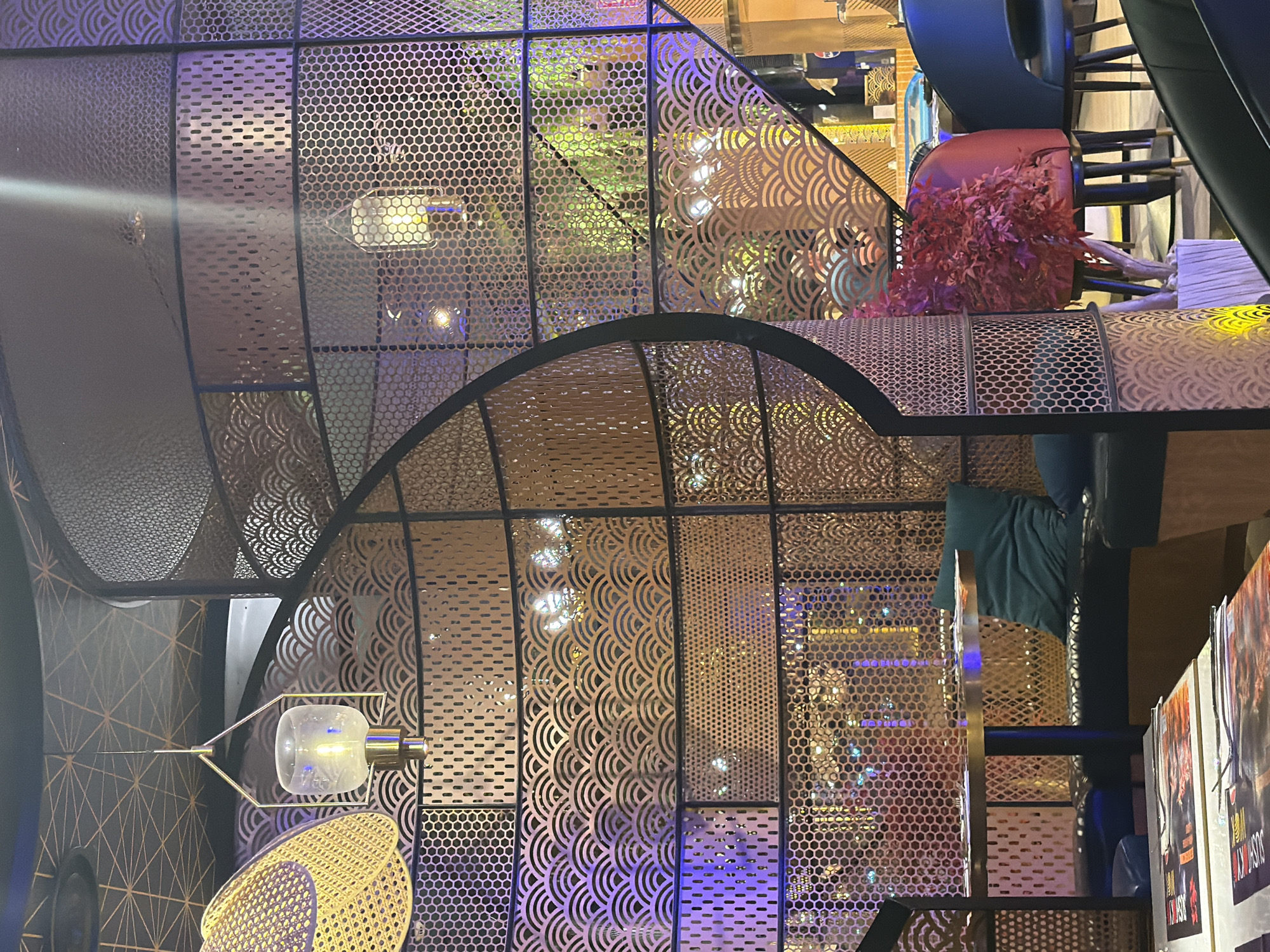 An interior scene depicts a dining area with ornate metallic partitions. A round table with a metallic top is visible, accompanied by dark cushioned seating and a teal pillow. In the foreground, two tables are set with menus and cutlery. The partitions feature geometric and wave-like patterns, illuminated by various colored lights, including blue and purple. Hanging lamps with glass shades and exposed bulbs add to the ambiance. To the right, a red upholstered chair is adorned with a bushy red plant. In the background, other diners and tables are partially visible through the perforated screens, along with hints of foliage.