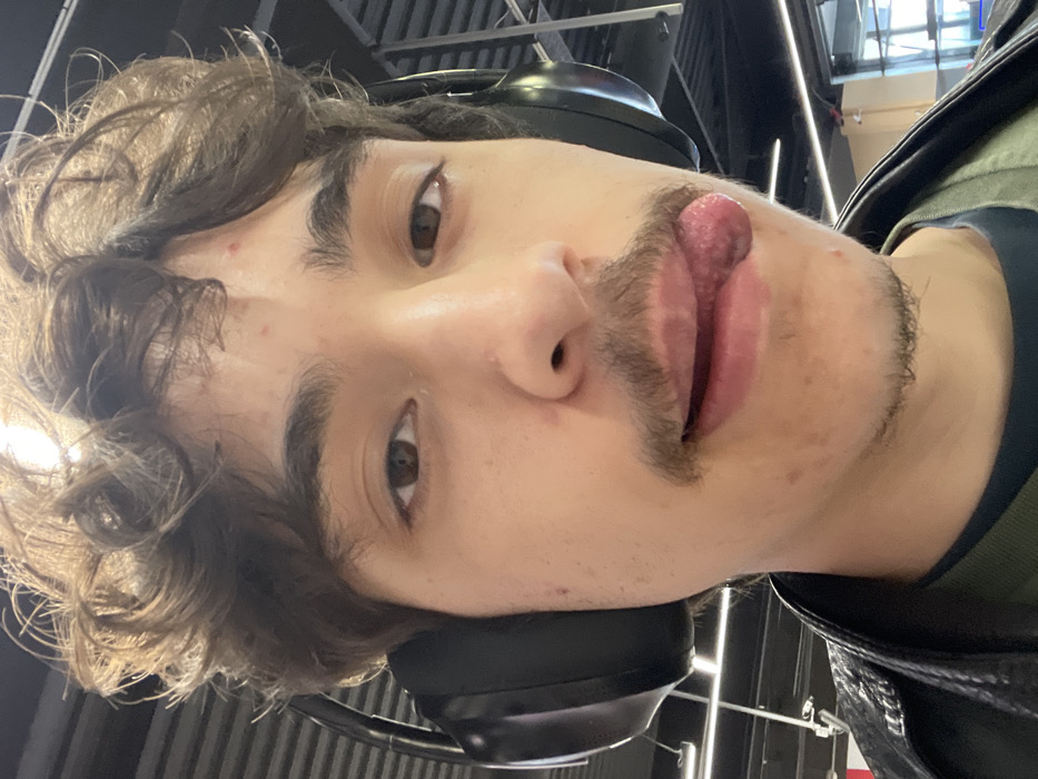 The image displays a close-up, frontal portrait of a young male individual, approximately 18-30 years old, situated indoors in Puyvert, France. He possesses curly brown hair, dark brown eyes, and a mustache. Several small red blemishes are visible on his forehead and cheeks. The individual is wearing black over-ear headphones, a dark-colored shirt, and a black leather-style jacket. His tongue is extended from the right side of his mouth, curving slightly downwards. The background is blurred, revealing an indoor setting with a high ceiling, horizontal dark paneling, and bright overhead lighting fixtures. A faint reflection on the right side suggests a window or glass partition.