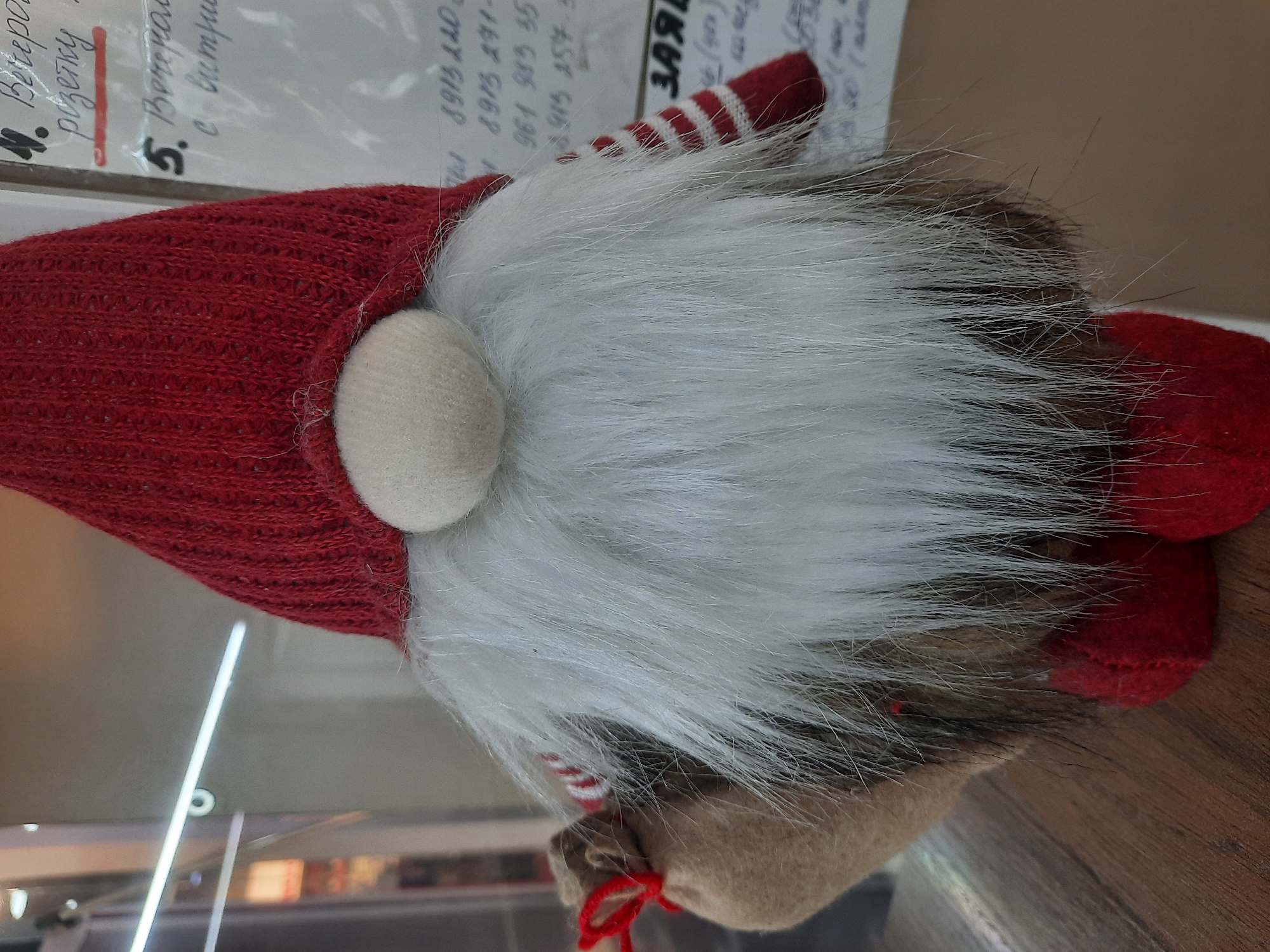 An image, rotated clockwise, features a decorative gnome figure positioned on a wooden surface, with paper documents in the background. The gnome wears a textured red knitted hat and has a prominent, fluffy white beard that extends over its body, with hints of darker brown/grey fur beneath. A small, round, beige nose protrudes from under its hat. The gnome's hands are visible as red mittens with white stripes, and it appears to be holding a small, light brown sack tied with a red string. Red feet are also visible at the base of the figure.
The background consists of several papers with handwritten and possibly printed Cyrillic text and numbers. Visible text includes "розетку" (socket/outlet), "5. Вечера" (evening), "с витрие" (from the display/window, or partial), "ЗАЯВ" (likely part of 'application'), and phone numbers such as "8913 220", "961 983 35", and "0915 157". Below the papers, a reflective surface, possibly glass or plastic, shows blurry reflections of indoor lighting and structures. The wooden surface beneath the gnome adds a warm, earthy tone to the setting. No specific activity is taking place; it is a static display of the decoration. The scene is indoors, likely in an office or retail environment given the paperwork and display-like qualities.
