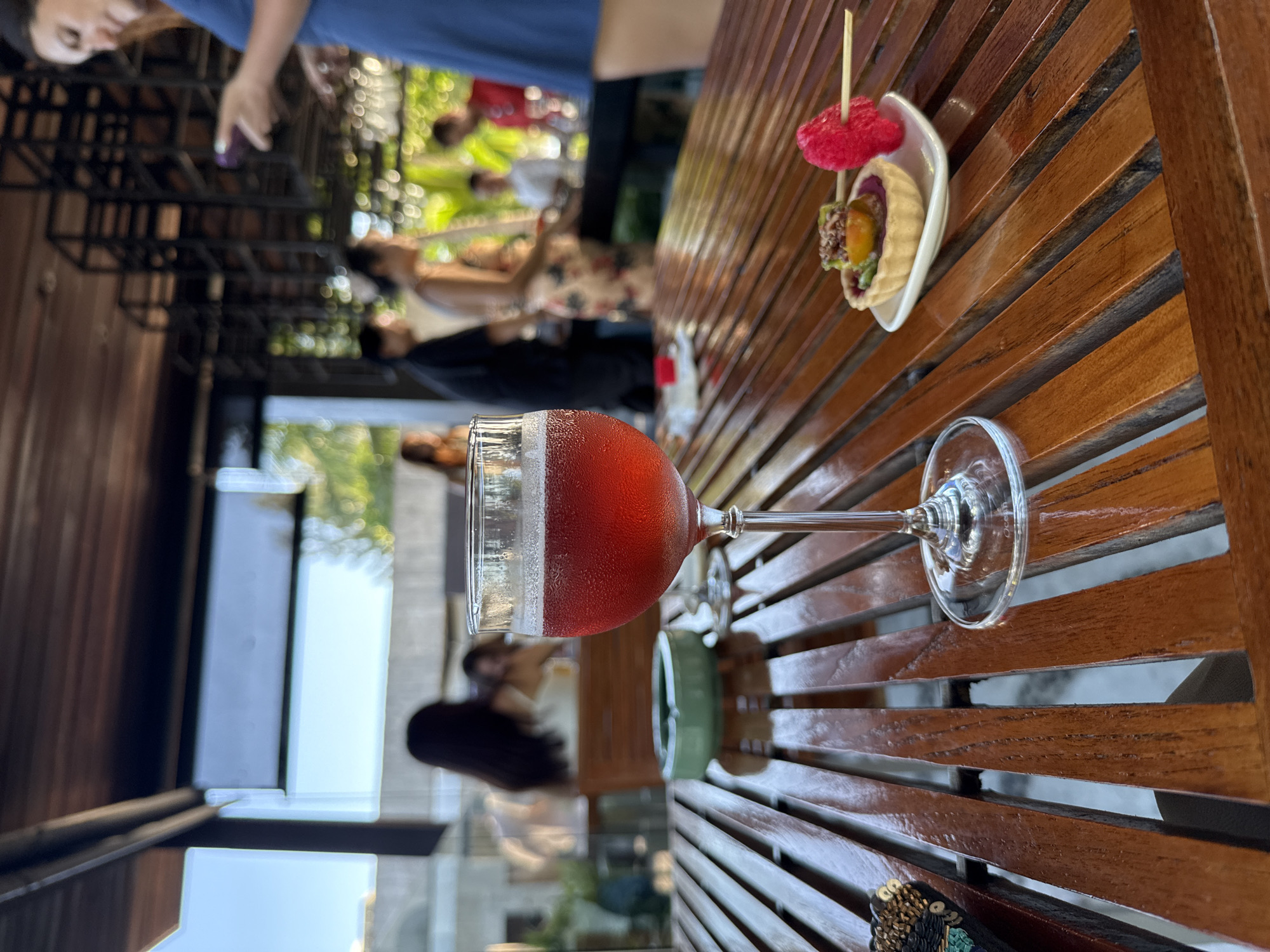 In Baan Bo Phut, Thailand, a close-up shot focuses on a glass of red beverage with condensation on a wooden slatted table. To the right, a small tart with purple filling and fruit toppings sits on a white saucer, adorned with a red, flower-shaped garnish on a toothpick. In the background, out of focus, several individuals are seen at tables and standing, suggesting a restaurant or bar setting with an open-air covered structure and greenery. A woman in a blue dress is partially visible on the far right.