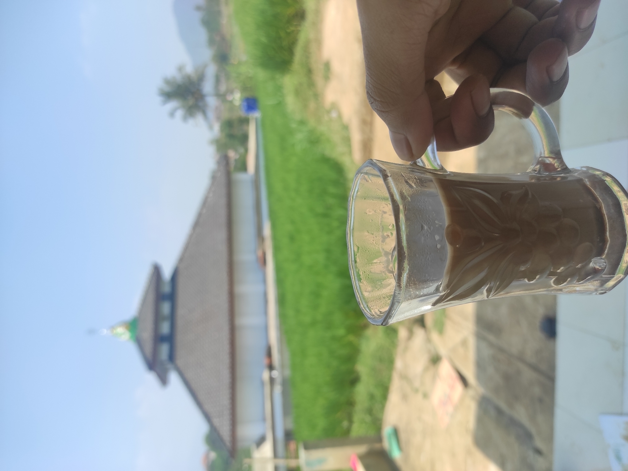 A first-person perspective shot shows a clear glass mug filled with a dark beverage, held by a person's hand. The mug is decorated with embossed floral patterns and is condensation-covered, suggesting it contains a cold drink. The background features a mosque with a grey tiled roof and a green-topped dome, set against a bright blue sky. In front of the mosque, a vibrant green field of crops, likely rice paddies, stretches across the landscape, characteristic of rural agricultural areas. A palm tree is visible to the right of the mosque, and distant hills complete the scenic view. The overall scene depicts a tranquil setting, possibly in a village or rural area near Sumedang, Indonesia, given the architectural style of the mosque and the lush, cultivated landscape.