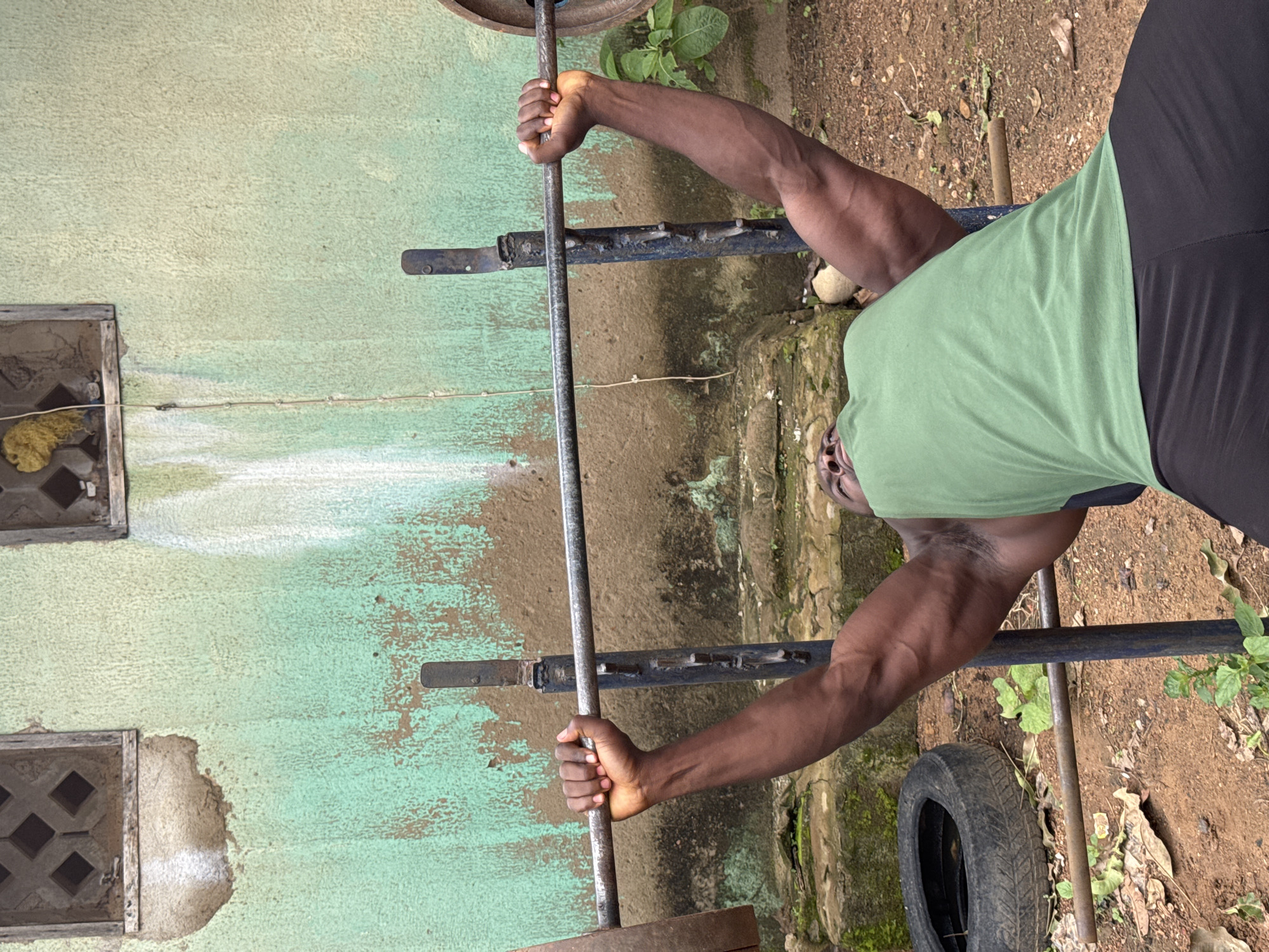 A man is performing a bench press outdoors in Akwanga, Nigeria. He is lying on his back on a weight bench with a barbell overhead, which is supported by a rack. He is wearing a green t-shirt and black pants. The background consists of a textured wall with two square openings and an overgrown, somewhat dilapidated outdoor space with dirt ground, rocks, and vegetation. A discarded tire is visible to the left of the bench. The lighting suggests it is daytime.