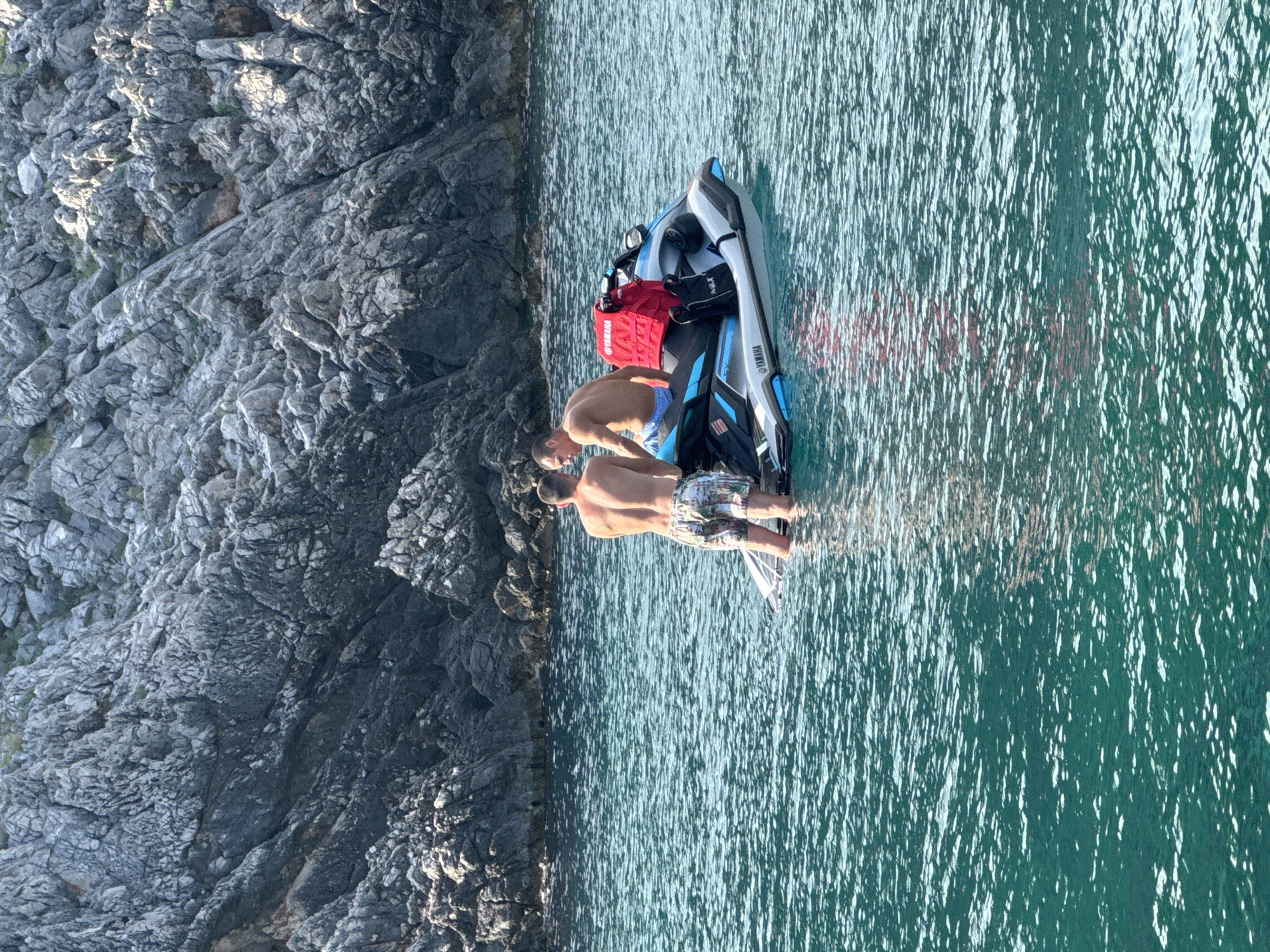 Two shirtless men are on a jet ski in the clear, turquoise waters near a rocky cliff face. One man, wearing patterned swim trunks, stands in the water next to the jet ski, facing away from the camera. The other man, wearing blue swim trunks, is seated on the jet ski, also facing away. A bright red life vest is attached to the back of the jet ski, along with a black bag. The jet ski has "YAMAHA" branding visible on its side. The rocky cliffs, characteristic of the coastline in Dubraljevina, Montenegro, form a backdrop to the scene. The water is calm with light ripples and reflections of the sun.