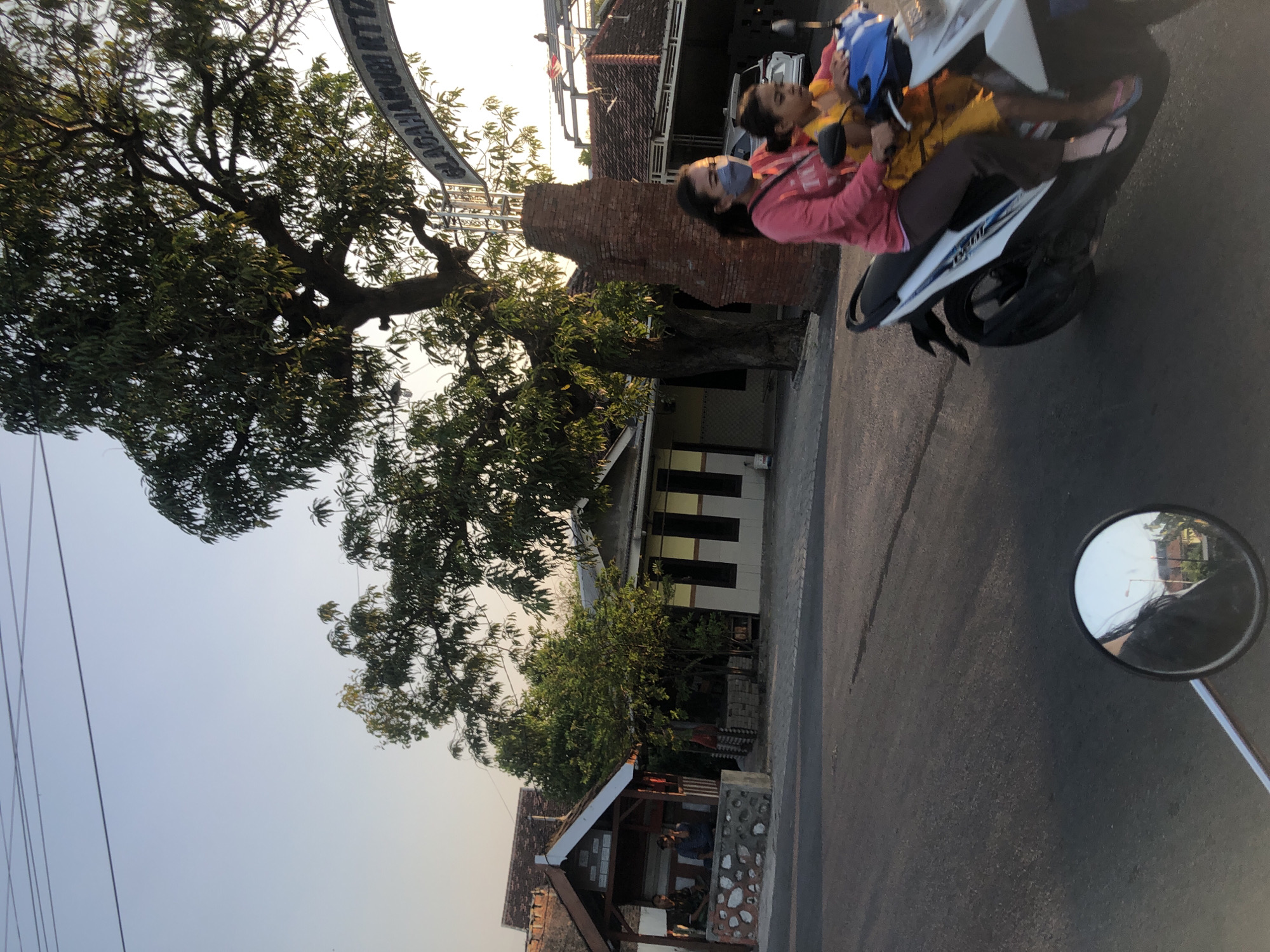The image depicts a street scene in Glagah Arum, Indonesia, during the late afternoon or early evening. The sky is clear and a pale blue, with power lines crisscrossing the upper portion of the frame. A large tree with dense foliage dominates the middle ground, casting shadows on the surrounding area. To the left of the tree, a small, open-sided structure with a wooden roof and stone base is visible, where two individuals are seated. Adjacent to this structure is a building with a yellow and gray facade and dark windows.
In the foreground, a white scooter is being ridden by two individuals. The person in front, presumably an adult woman wearing a pink long-sleeved shirt and brown pants, has a light blue mask covering her lower face. She is driving the scooter. The person behind her, a child wearing a yellow shirt, is seated in front of her. They are both wearing flip-flops. The scooter is moving on an asphalt road.
A brick pillar and an arched sign are visible to the right of the tree, with the text "GLAGAHARUM RTT" partially visible on the sign. Further in the background, a white car is parked and a tiled roof of a building is visible. A motorcycle's circular rearview mirror is in the bottom left corner of the image, reflecting a street lamp and buildings. The overall impression is a snapshot of everyday life in a residential or semi-commercial area of Glagah Arum.