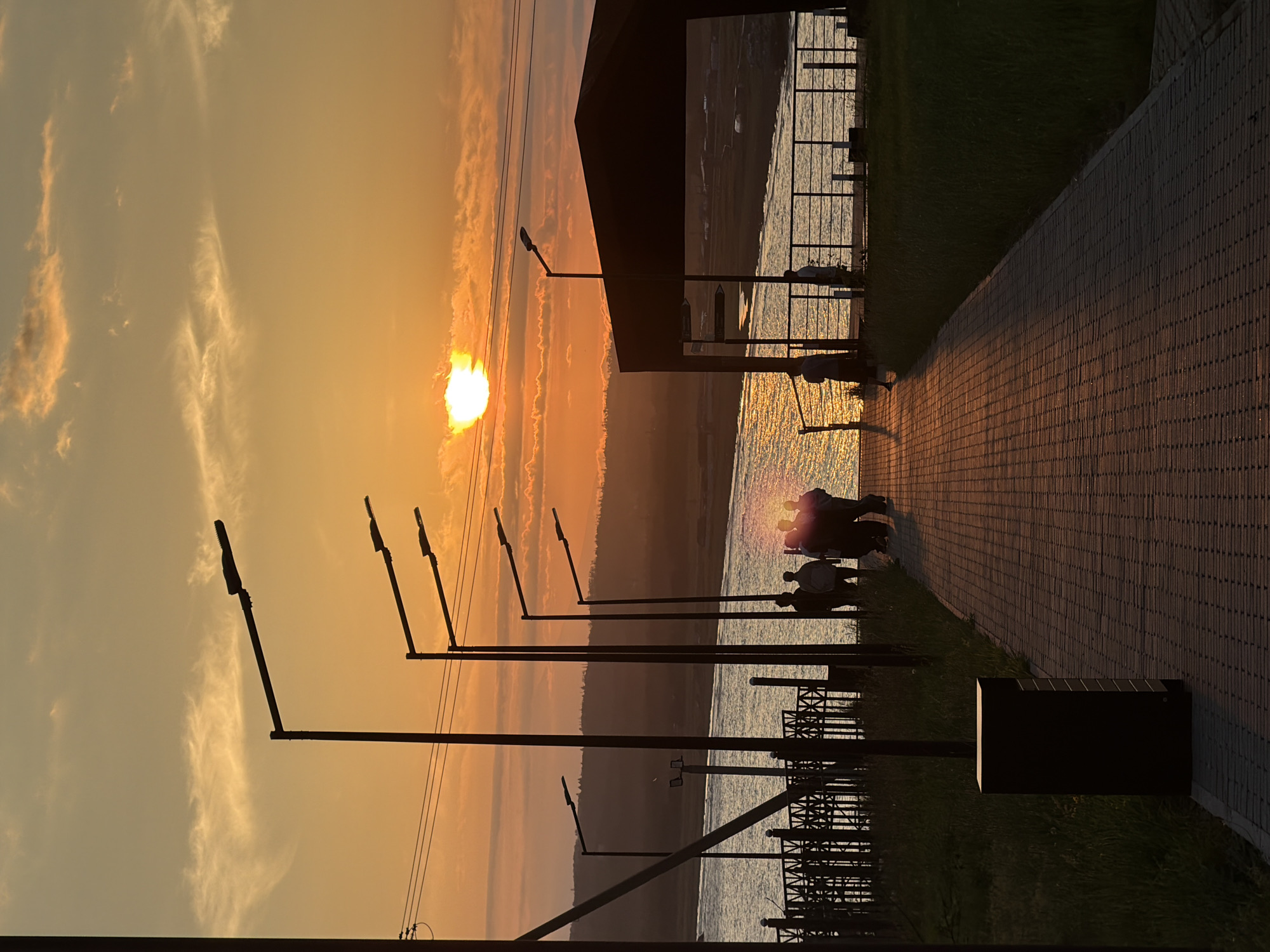 The image depicts a sunset scene in Yuzhno-Kurilsk, Russia, likely along a waterfront promenade. The sky is ablaze with the warm hues of orange and yellow, with scattered clouds catching the last rays of the sun. The sun itself is a bright, glowing orb partially obscured by the horizon, casting a shimmering reflection on the calm water.
In the foreground, a paved walkway leads towards the water. Several lampposts with modern designs are spaced along the path, silhouetted against the bright sky. To the right, a covered structure, possibly a gazebo or pavilion, stands with its dark roof creating a strong contrast with the sunset.
A group of people are visible in silhouette on the promenade near the water's edge. They appear to be casually gathered, with some facing the water and others interacting amongst themselves. Further down the promenade, another individual is seen walking. The ground on either side of the walkway is covered with green grass. In the middle ground, a body of water, likely the Sea of Okhotsk, stretches out, reflecting the vibrant colors of the sky. Beyond the water, a distant shoreline with rolling hills can be observed, also bathed in the warm light of the sunset. The overall atmosphere is serene and tranquil, capturing the beauty of a coastal sunset.